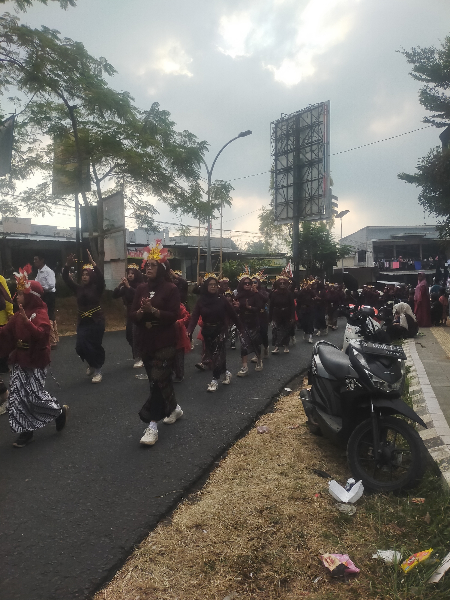 A group of women in matching dark maroon tops and patterned skirts are parading down a paved street in what appears to be a village in Tuwel, Indonesia. They are wearing decorative headpieces with feathers and what look like crowns. Some of the women have their hands raised in a synchronized gesture. A motorbike is parked on the side of the road next to a grassy area with some scattered litter. In the background, there are buildings, trees, and a large billboard. The sky is overcast with some patches of sunlight breaking through. The overall scene depicts a cultural procession or celebration.