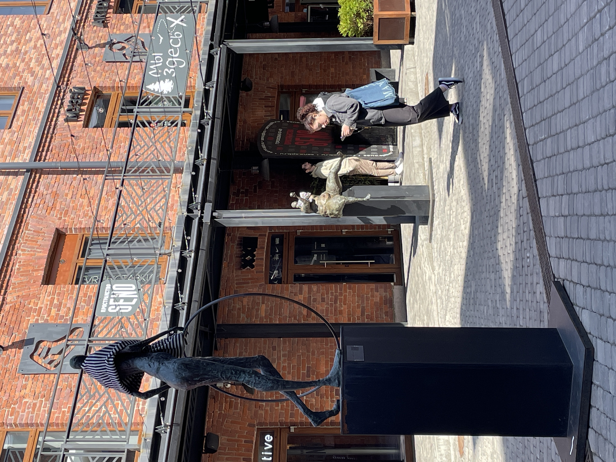 Here is a description of the image:
The image captures a street scene in Saint Petersburg, Russia, displaying an urban architectural composition. Dominating the foreground is a modern sculpture of a figure appearing to levitate within a large hoop, standing atop a black pedestal. The architecture in the background consists of brick buildings with visible metal structural elements. A person is positioned mid-frame, appearing to use a mobile phone, while a second sculpture is visible further behind. The location seems to be a pedestrian zone or courtyard with cobblestone pavement. Signage written in Russian is present, adding to the regional context.