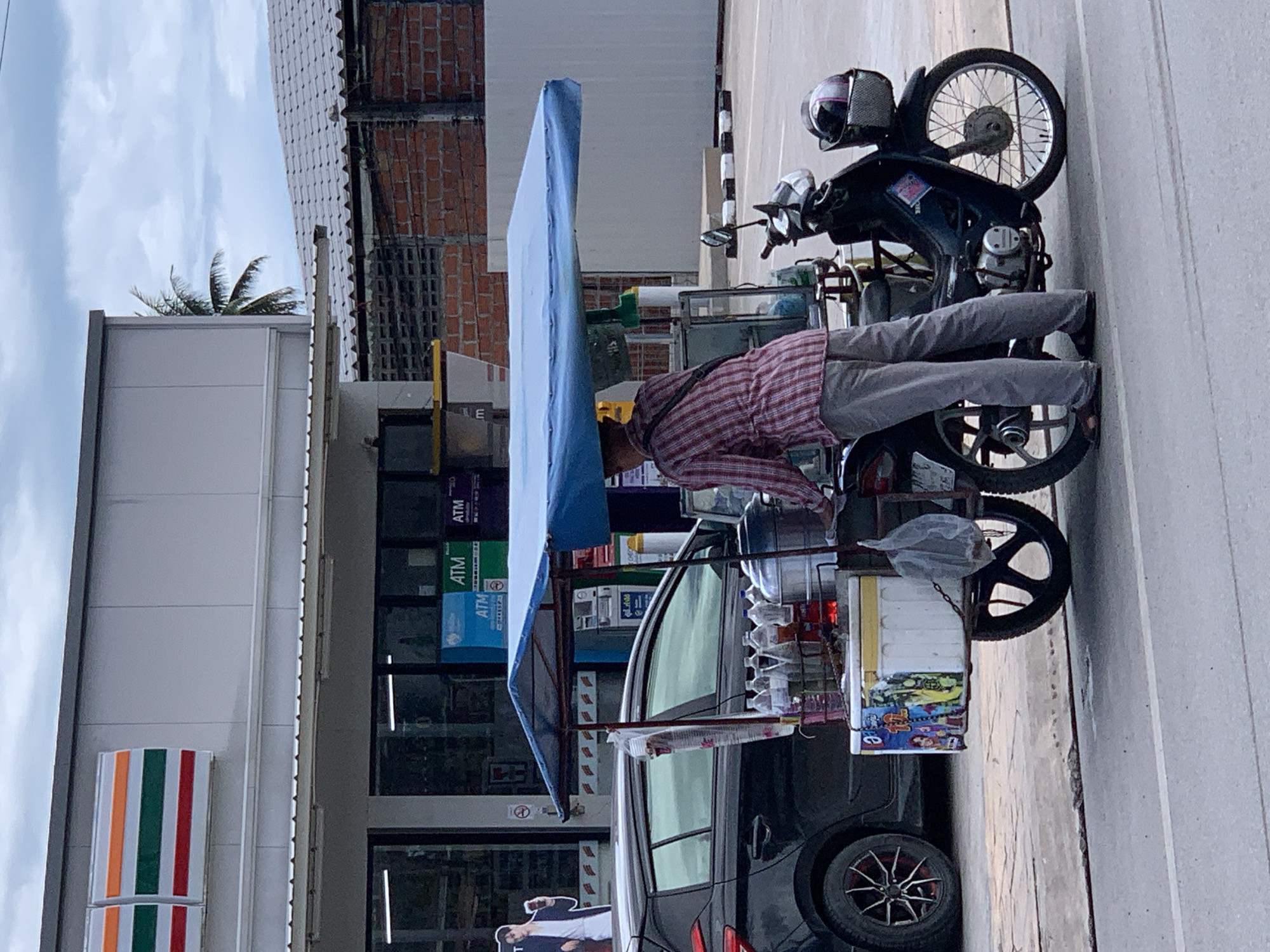 This image depicts a street vendor operating a mobile food cart attached to a motor scooter in Baan Bo Phut, Thailand, positioned on the shoulder of a paved road in front of a 7-Eleven convenience store.
A single individual, a street vendor, is seen from the back, wearing a red and white plaid shirt, gray trousers, and sandals. The vendor is standing beside a blue-canopied mobile cart, which is affixed to a dark-colored Honda motor scooter. The cart contains a collection of clear plastic bottles, a stack of clear plastic cups, a large metallic steamer pot, and a white insulated cooler box decorated with colorful imagery. A clear plastic bag hangs from the cart's side. The motor scooter has a helmet in its front basket and a partially obscured license plate on the rear fender.
In the background, the front of a 7-Eleven store is visible, featuring its characteristic orange, green, and red striped logo. Multiple ATM machines (blue and green) are discernible through the store's large glass windows. To the left of the store's entrance, a life-size promotional cutout of a man holding a drink cup is displayed. A black hatchback car is parked between the vendor's cart and the 7-Eleven building. Beyond the immediate commercial structures, a low brick wall and a building with a corrugated metal roof are visible, along with a palm tree under an overcast sky. The foreground shows a light gray asphalt road with a single white line.