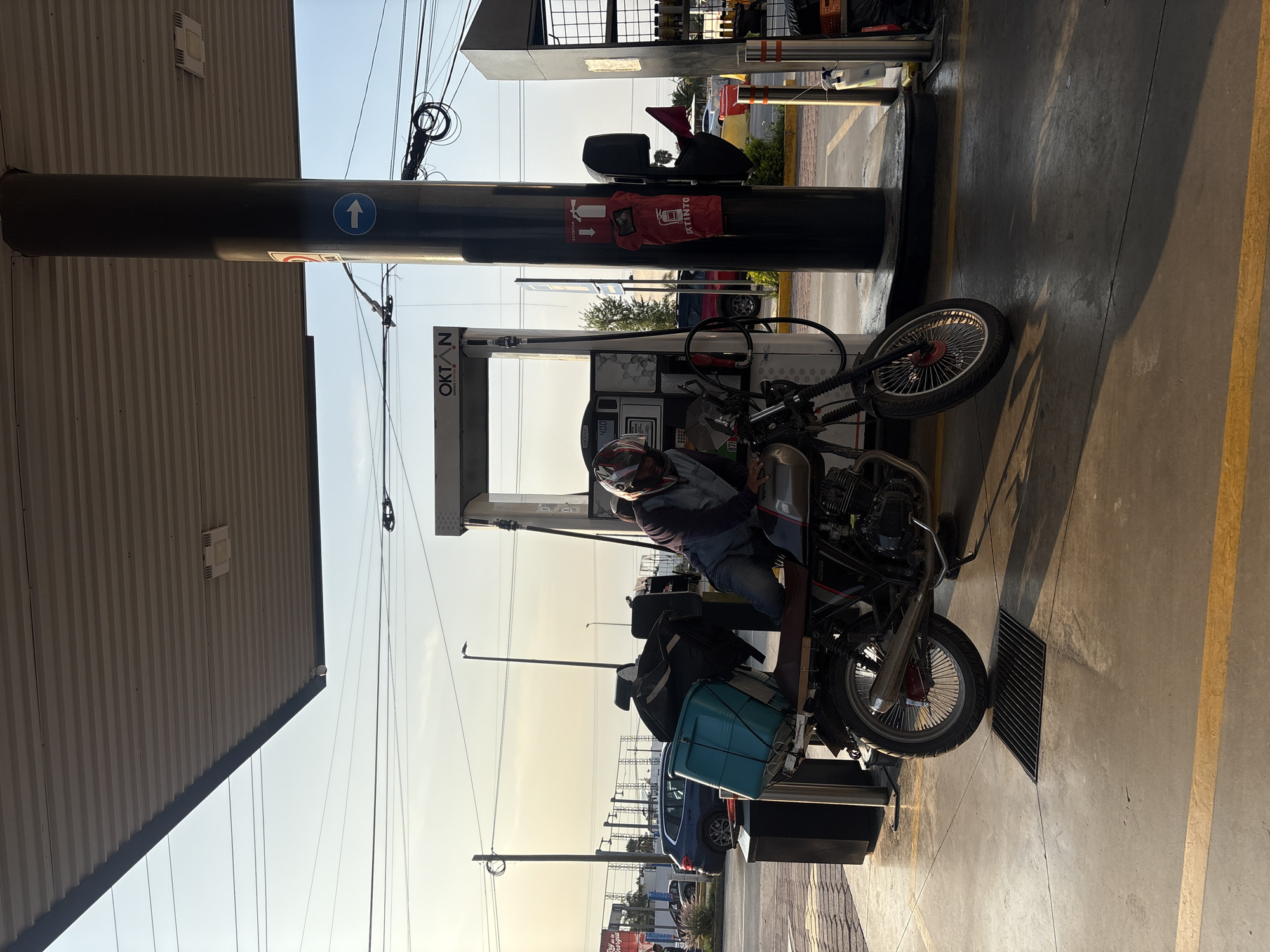 This landscape image depicts a person refueling a motorcycle at a gas station in San Bernardino Tlaxcalancingo, Mexico. The setting appears to be late afternoon or early morning, characterized by long shadows on the concrete ground and a clear, bright sky with a warm, soft glow.
The main subject is a person seated on a black motorcycle with spoked wheels, positioned beside an "OKTAN" branded fuel pump. The person is wearing a helmet, a denim jacket, and jeans, and appears to be in the process of fueling the motorcycle, as the hose is connected. The motorcycle is loaded with luggage, including a black bag and a light blue/teal container strapped to its rear, suggesting a journey.
A prominent dark grey pillar, supporting the gas station canopy, stands on the left. Attached to this pillar are several signs: a blue circular sign with a white upward arrow, and a red fabric cover or bag clearly labeled "EXTINTOR" (Spanish for extinguisher) alongside an image of a fire extinguisher, indicating the presence of safety equipment. The fuel pump's digital display shows numbers, including "4.00," likely indicating price or volume.
In the background, a blue car is parked, and other vehicles are visible in the distance. The gas station structure features a corrugated metal roof section. Numerous power lines and cables are visible against the clear sky. The concrete ground includes yellow painted lines and a metal grate or drain cover. The scene captures a routine stop at a service station under good weather conditions.