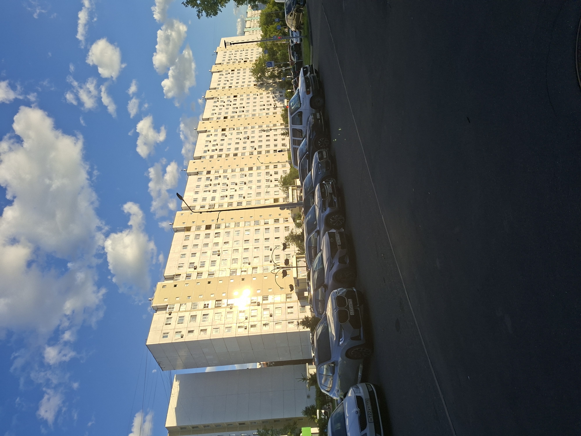 The image shows a long, multi-story apartment building with a light-colored facade and numerous windows. The building appears to be residential, with air conditioning units visible outside many windows. In front of the building, a line of cars is parked along a street. The cars are varied in make and model, with some appearing to be SUVs and sedans.
The scene is set against a bright blue sky with scattered white clouds, suggesting fair weather. Sunlight reflects off the windows of the apartment building, indicating it is daytime, likely late afternoon or early evening due to the warm glow. Trees are visible behind the cars and to the left of the building, suggesting an urban or suburban setting with some greenery.
The image is oriented vertically, with the building and sky occupying the left side and the street and cars on the right. The perspective is from a slightly elevated viewpoint looking down at the parked cars and across at the building. There is no discernible activity or event taking place other than the static scene of parked cars and the building.
Visible text on license plates includes "H122XT190" and "6035 HPA".
The overall impression is of a typical urban residential area in Moscow, Russia, on a clear day. The image is factual and objective, with no overt emotional content.