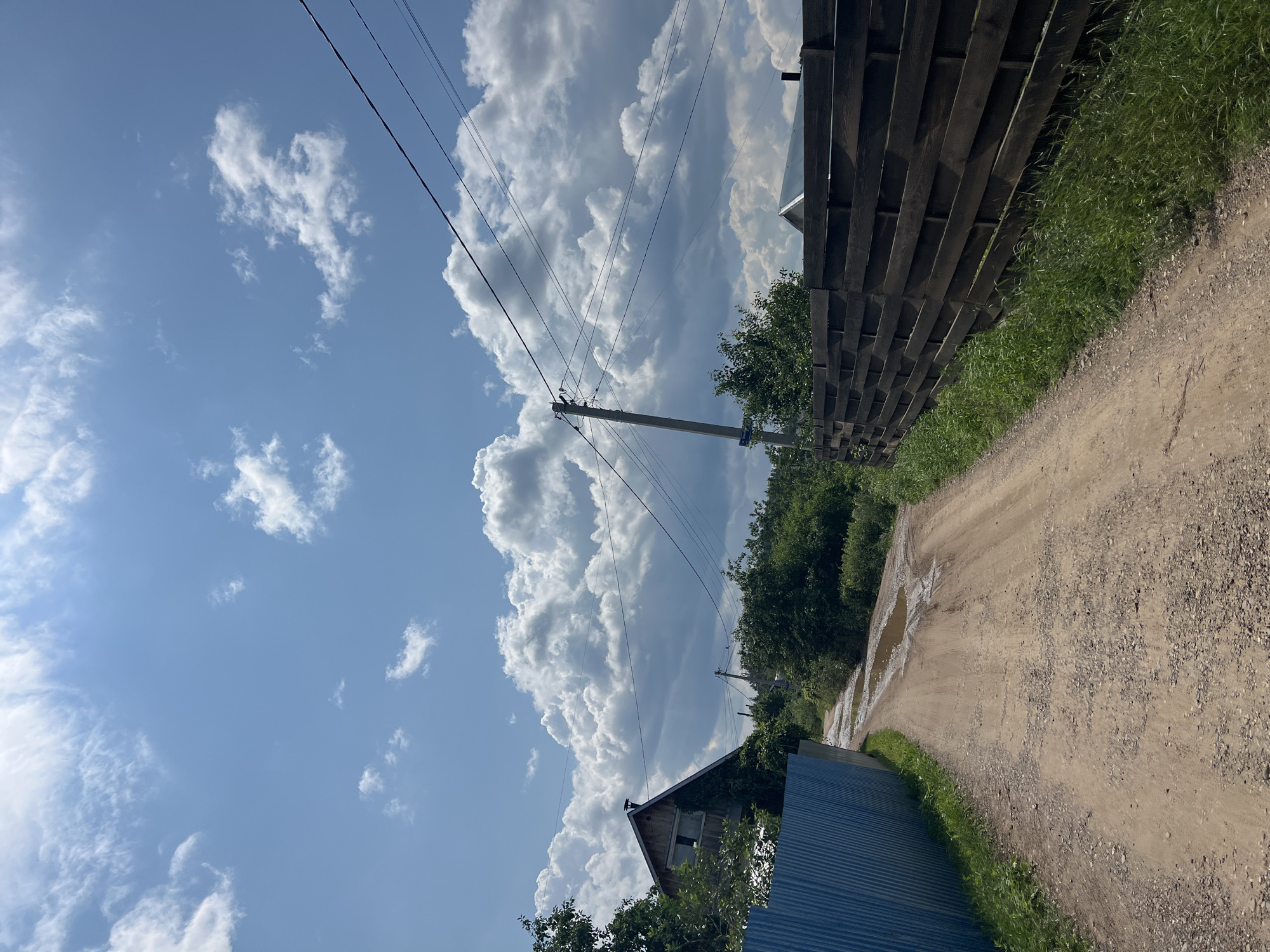 The image depicts a dirt road in Noviy, Russia, on a cloudy day. The road is lined with a blue metal fence on the left and a dark wooden fence on the right. Green trees and bushes flank the road, and a wooden house with a gabled roof is visible on the left side of the image. A utility pole with electrical wires crosses the sky. The road has several puddles, indicating recent rainfall. The sky is bright blue with scattered white clouds, suggesting it is daytime.