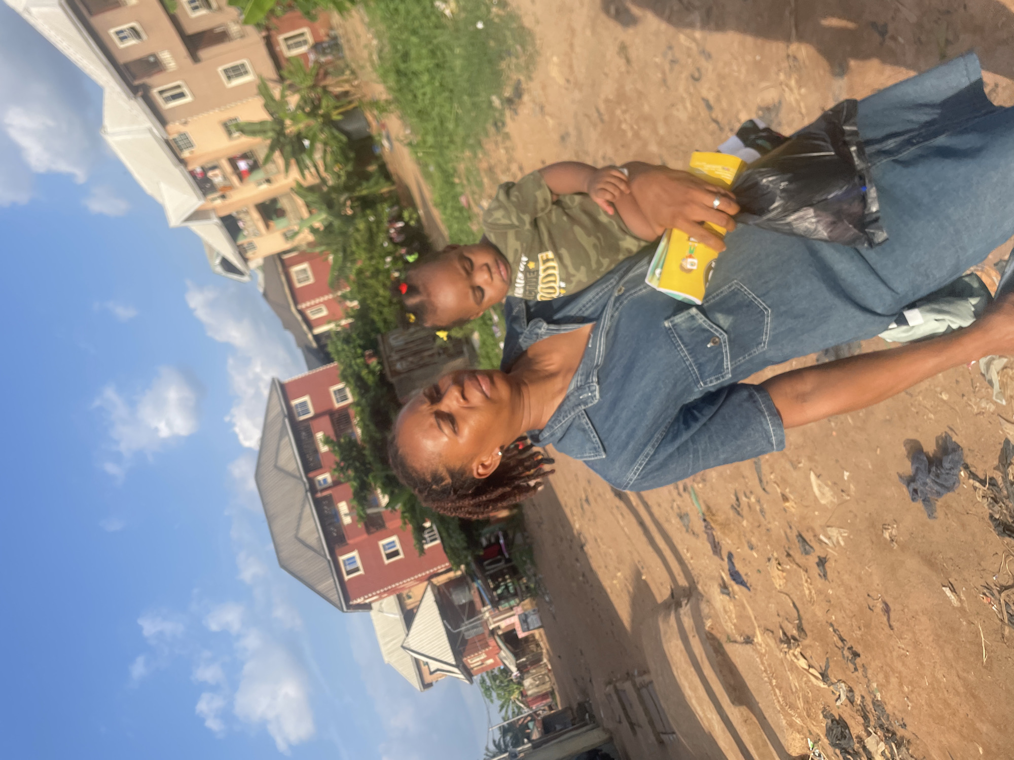 This image, taken in Nkpor, Nigeria, captures a candid outdoor moment featuring a woman and a young child under a bright, sunny sky. The photo is slightly rotated, showing the subjects and background at an angle.
The main subjects are a woman and a small child. The woman, with dark skin and dark dreadlocks, is positioned in the foreground, looking slightly off-camera with a calm, pleasant expression and a subtle smile. She is wearing a blue denim short-sleeved dress. Her right hand, adorned with a silver-colored ring, holds a stack of yellow items, possibly books or product packaging, and a black plastic bag. One of the yellow items shows a colorful cartoon character.
A young child, likely a toddler, is standing in front of the woman, held by her. The child is wearing a camouflage-patterned t-shirt with the partial word "CTIVE" visible on it. The child's hair is styled in small braids with colorful accessories, and they are looking towards the camera with a neutral expression.
The setting is an unpaved, reddish-brown dirt area, possibly a street or open ground, with some scattered debris and sparse green vegetation. In the background, several multi-story residential or commercial buildings with red, beige, and brown facades are visible. The sky is clear and bright blue with scattered white clouds, indicating it is daytime, likely morning or afternoon.
