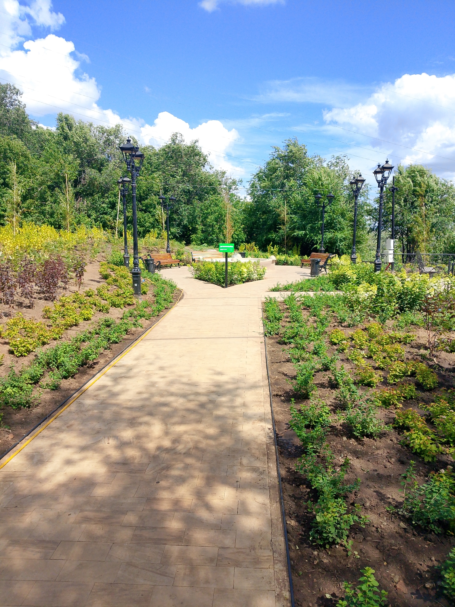 The image depicts a park pathway in Orenburg, Russia, on a sunny day. A paved pathway, made of light brown square tiles, leads into the distance, flanked by meticulously landscaped gardens. On either side of the path, there are beds of young plants with green and reddish-purple foliage, indicating recent planting or active garden maintenance. In the background, there are several vintage-style black lampposts with multiple lights. Wooden benches and trash receptacles are visible near the lampposts, suggesting a public space designed for relaxation and recreation. Beyond the gardens and pathway, a dense line of lush green trees forms the backdrop, suggesting a wooded area or a larger park. The sky is a vibrant blue, dotted with fluffy white clouds. A green sign with white text, possibly indicating park rules or directions, is positioned on the right side of the pathway. The scene appears serene and well-maintained, a typical urban park setting.