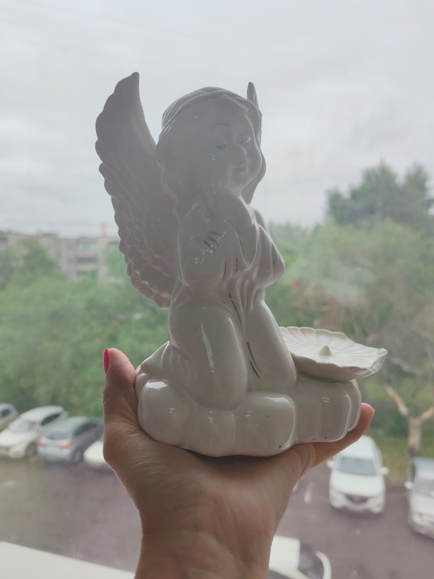 A white porcelain figurine of a cherub or angel is held in a person's hand. The figurine is seated on a cloud, with its wings spread behind it. It holds a small, shell-like dish with a tiny point in the center. The figurine has a serene expression. The background is a blurry view through a window. Outside, trees and buildings are visible, and several cars are parked in what appears to be a residential area. The sky is overcast, suggesting a cloudy day. The person's hand holding the figurine has brightly painted pink fingernails. The overall lighting is soft and diffused. The image appears to be taken indoors, looking out. The location context is Komsomolsk-on-Amur, Russia. No visible text is present in the image.