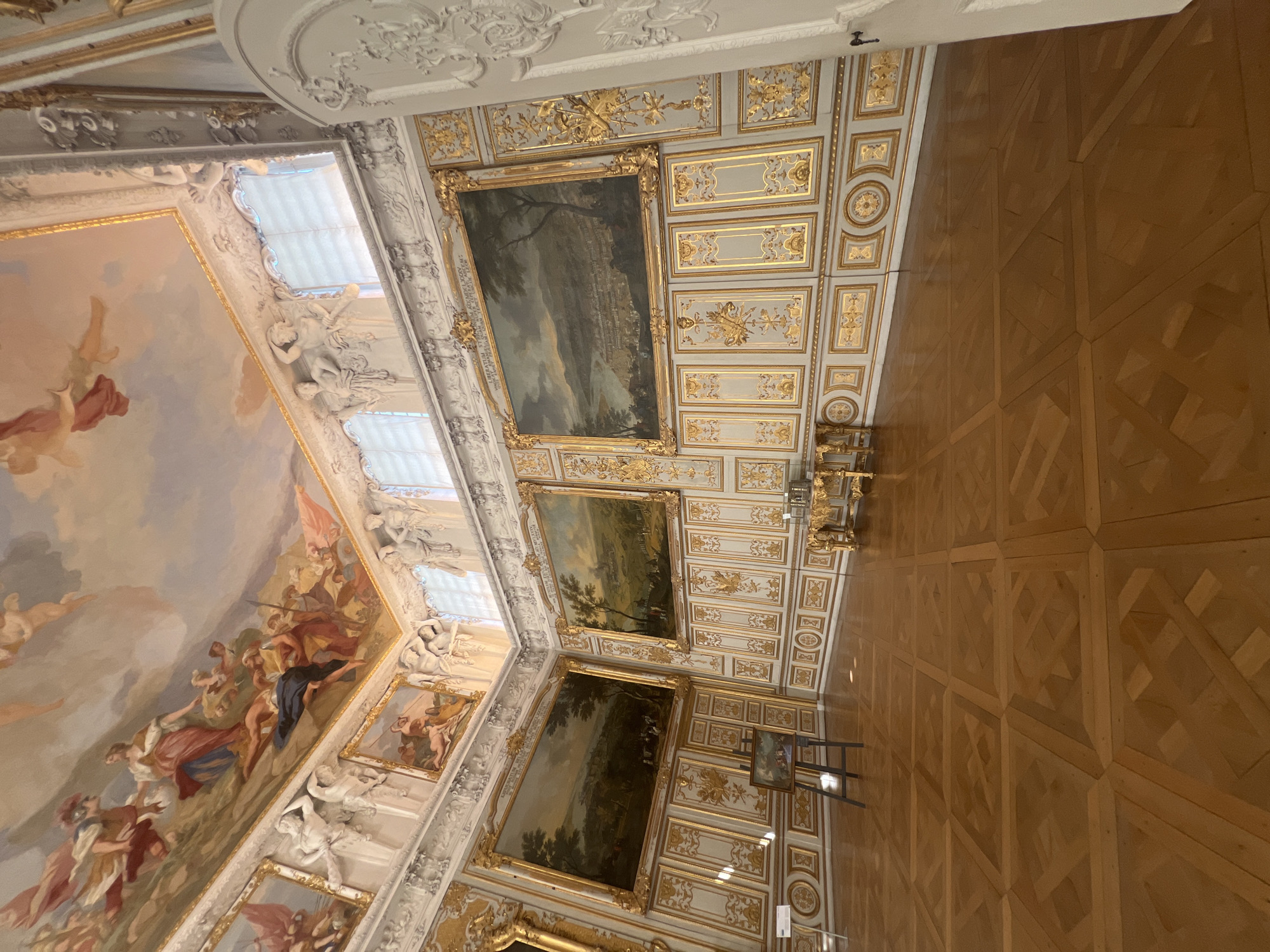 The image shows the interior of a richly decorated room, likely a palace hall, in Oberschleißheim, Germany. The room features ornate walls with gold detailing and large framed paintings. The ceiling is adorned with a fresco depicting mythological or celestial scenes with figures. The floor is a polished wooden parquet in a geometric pattern. In the room, there are several large landscape paintings on the walls. One painting is displayed on an easel in the foreground. There is also a decorative golden table and a white door on the right side of the image. The lighting suggests daylight entering from windows that are covered by blinds. There are no people visible in the scene. The overall impression is one of opulence and historical significance.