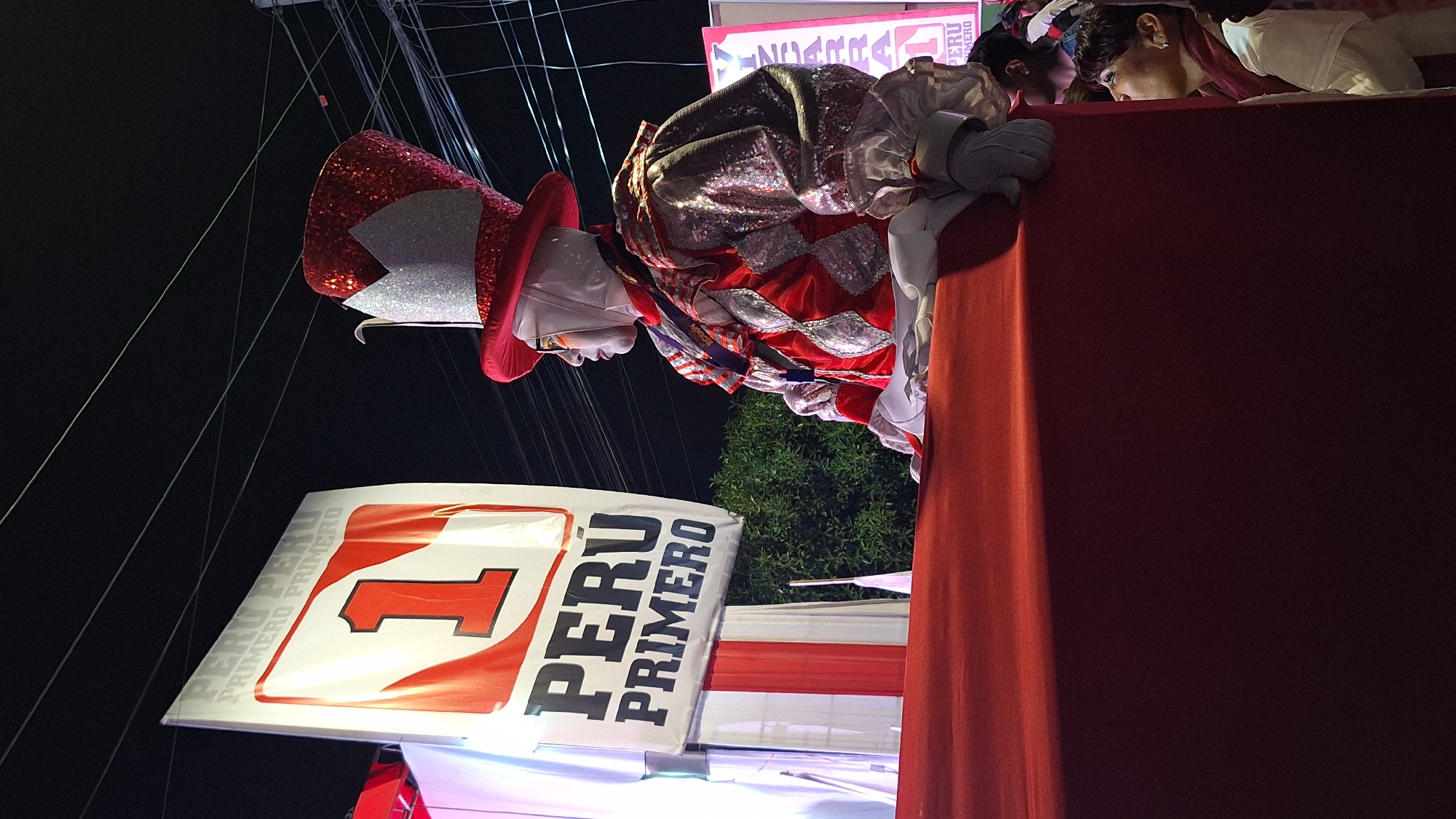 Captured at night in an unknown city, the image portrays a vibrant political rally or event. Prominently displayed is a banner with the words "PERÚ PRIMERO" and a bold red number 1, indicating a political campaign or slogan. Adjacent to the banner is a person dressed in a sparkly red and white costume with a matching top hat, likely a performer or supporter. A group of people are partially visible behind a red structure, potentially observing the event. Above, a web of power lines crisscrosses the dark sky. Vegetation, likely a tree or shrub, is visible in the background.