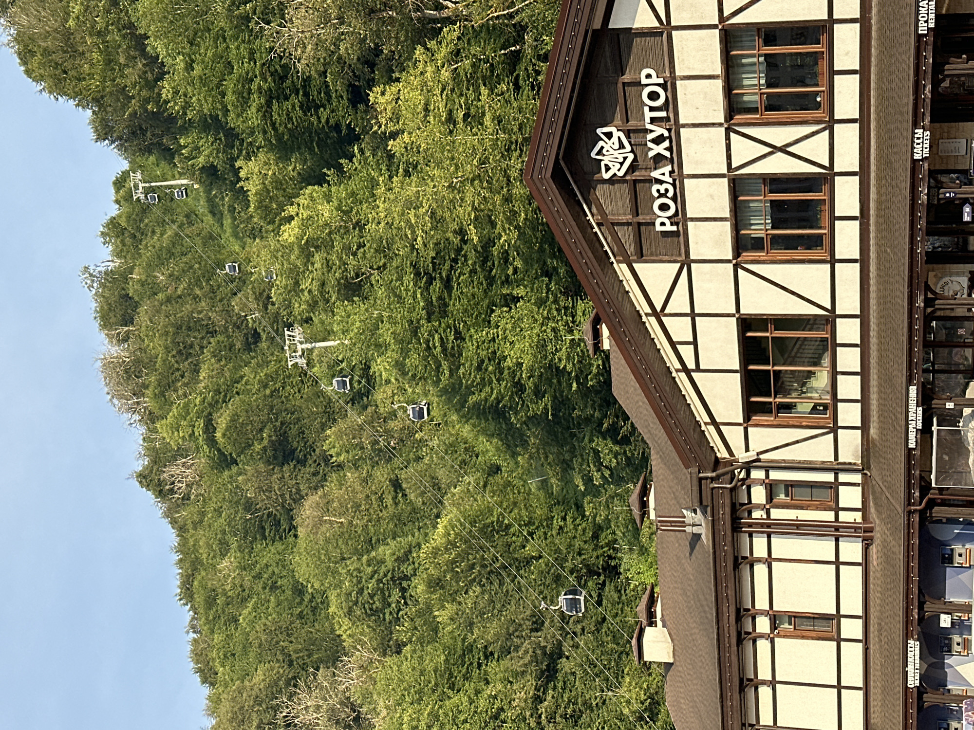 In the city of Estosadok, Russia, a mountain resort area, a picturesque scene unfolds under a clear blue sky. The foreground is dominated by a charming, timber-framed building, its upper facade adorned with the stylized text "РОЗА ХУТОР" (Rosa Khutor) and a geometric logo. This building appears to be a commercial establishment, with the lower level featuring a row of businesses, indicated by signage such as "КАССЫ TICKETS" (TICKETS) and "ПРОКАТ RENTAL" (RENTAL). The architecture suggests a blend of traditional and modern styles, with dark brown wooden beams contrasting against a light-colored stucco finish.
Rising behind the building is a densely forested mountainside, teeming with lush green trees. The vibrant foliage indicates a temperate climate, likely during the warmer months. Stretching across the mountainside, a series of ski lifts with multiple gondolas ascend the slopes. These gondolas, suspended by thin cables, are a prominent feature, illustrating the resort's recreational offerings. The ski lift towers are visible, strategically placed to support the aerial transportation system. The overall impression is one of a well-developed mountain resort, integrating natural beauty with modern infrastructure for tourism and leisure.