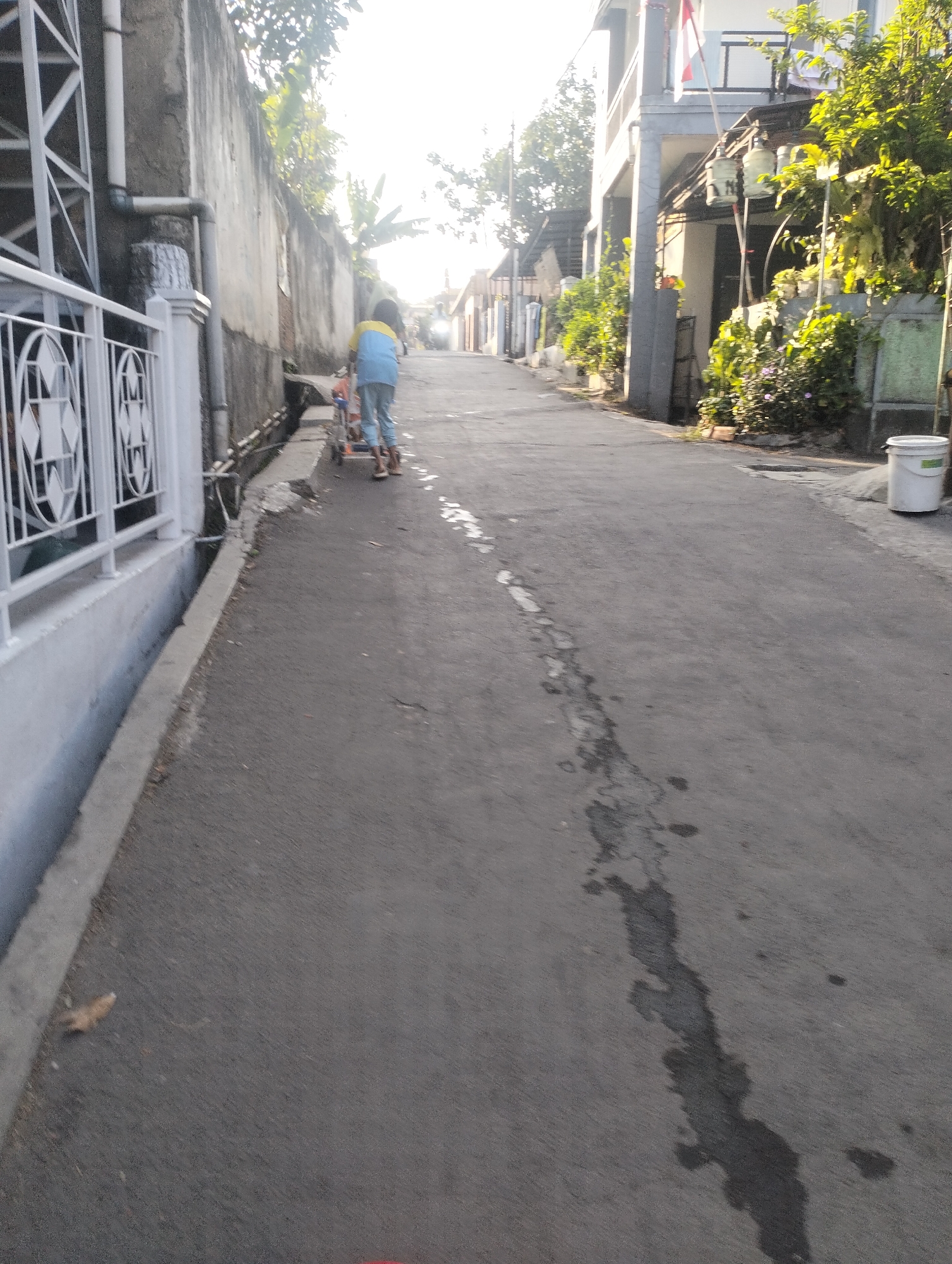The image depicts a narrow asphalt street in the Kel. Pasir Jati, Indonesia. A person, likely a child due to their size and attire, is seen from behind, pushing a small red and blue stroller. They are wearing a blue shirt with yellow accents and light-colored pants. The street is lined with residential buildings and a concrete wall. Lush green vegetation is visible on the right side of the street, bordering the buildings. Sunlight filters through the trees, casting shadows and creating a bright, somewhat overexposed atmosphere. A white plastic bucket is placed on the side of the road near the buildings. The road surface shows some wear and tear, with dark patches and cracks.