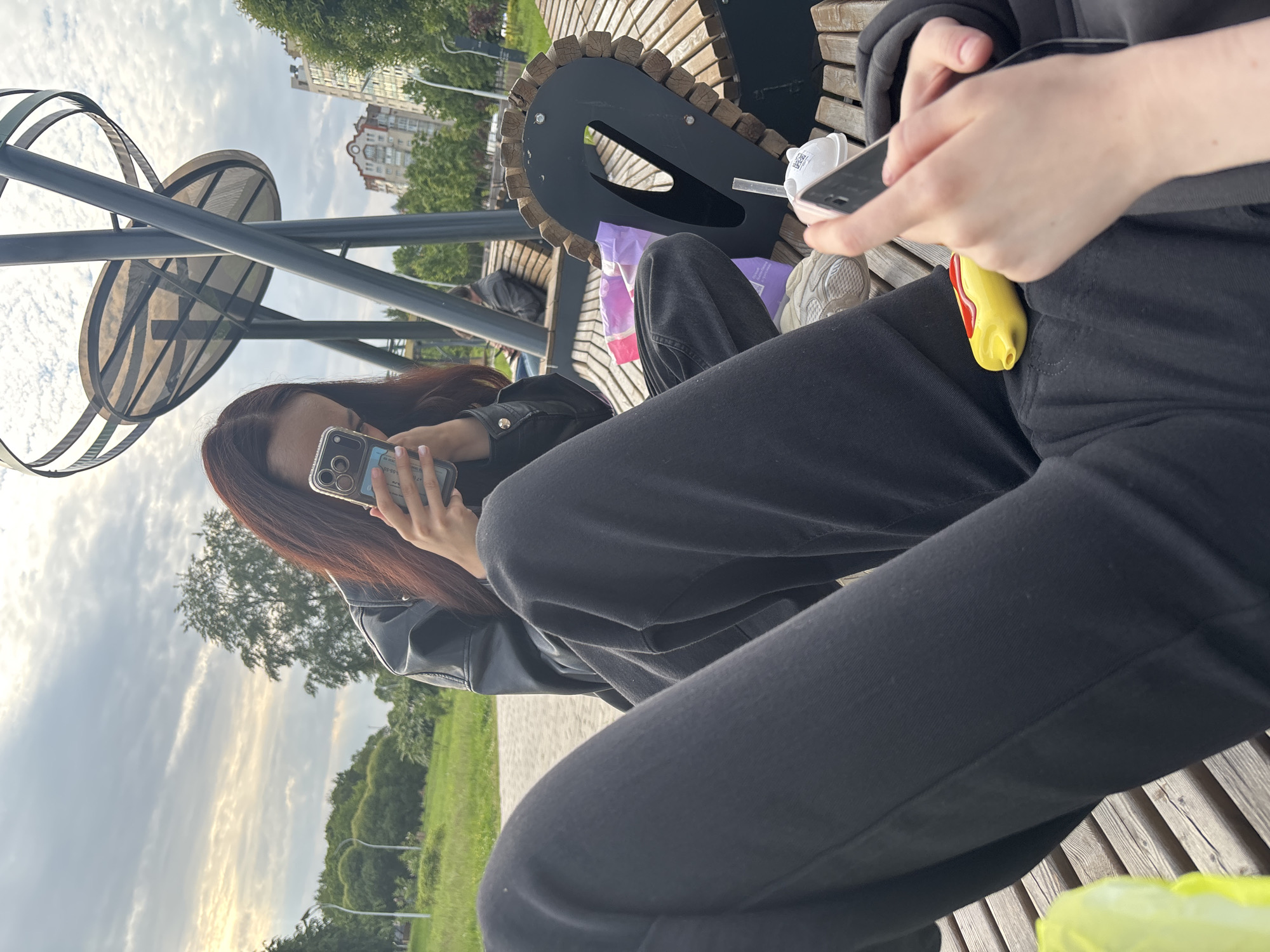 Two individuals are seated on a wooden park bench in Sinyavino, Russia. The person closer to the foreground is wearing black pants and a dark hoodie, holding a smartphone in their right hand. A yellow and red hot dog-shaped object rests on their lap. To their left, a lime-green plastic bag is partially visible.
The second person, positioned further back and slightly to the left, wears a black leather jacket and dark pants. They are holding a smartphone in both hands, seemingly taking a photo or recording a video. A portion of their dark hair is visible.
The park setting features trees, grass, and a paved pathway in the background. A distinctive metal sculpture with curved elements stands behind the individuals. Buildings can be seen in the distance. The sky is cloudy. Other park amenities, including a trash receptacle and plastic bags, are present on the bench.