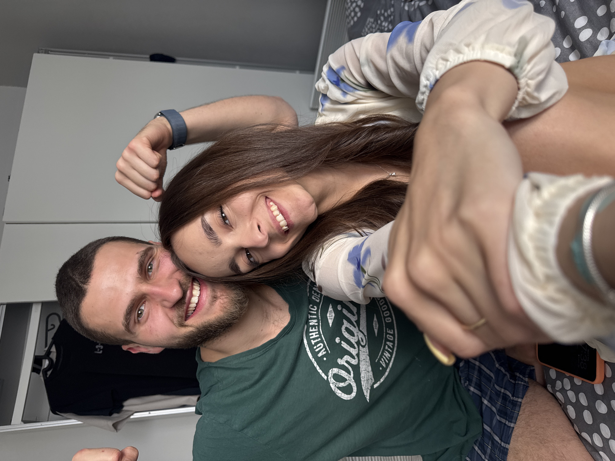 Two people are photographed in a close, intimate pose. A male with a dark beard and a green t-shirt is positioned on the left, while a female with long brown hair and a white floral top leans on him. They both smile at the camera. The environment appears to be an indoor space with a bed and closet visible in the background. Based on the subjects' clothing and pose, the location is a domestic setting. There is no information about the location in the city of Sofia, Bulgaria.