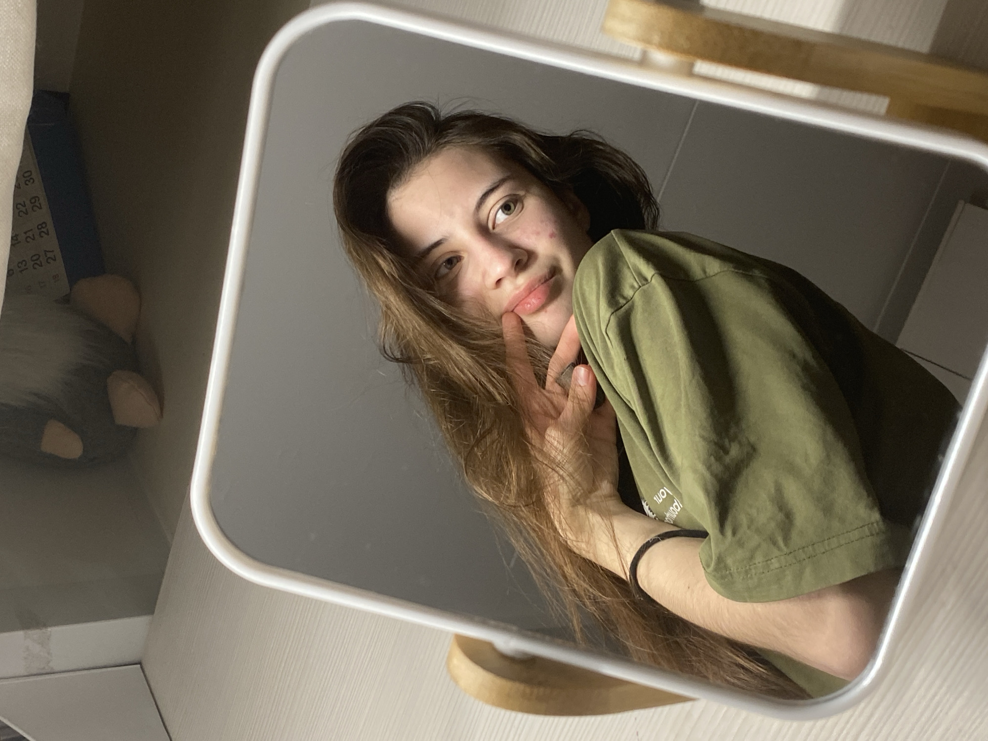 A young woman in Votkinsk, Russia, is depicted taking a mirror selfie in an indoor setting. The central subject is her reflection in a rectangular mirror with a white frame and rounded corners, positioned on a light-colored wooden surface.
The woman has long, brown hair and wears an olive green t-shirt. Her right hand is raised, with her index finger gently touching her lips, creating a thoughtful or slightly playful expression as she looks directly into the mirror. Some blemishes are visible on her skin, particularly on her right cheek. A black elastic band is worn on her left wrist.
Outside the mirror's frame, to the left, a cluttered background includes a piece of paper with printed numbers such as "13", "14", "15", "16", "17", "18", "20", "21", "22", "27", "28", "29", and "30," suggesting a calendar or measurement scale. A blue object and a fuzzy grey and white item, possibly a plush toy, are also visible. The reflected background within the mirror shows a plain dark grey wall and the continuation of the light wooden surface.
The scene is lit by indoor artificial light, giving it a soft, possibly evening ambiance. Visible white text on her olive green t-shirt, reflected backward, includes the word "long" and other partially obscured words that appear to read "iound!" and "mind!".