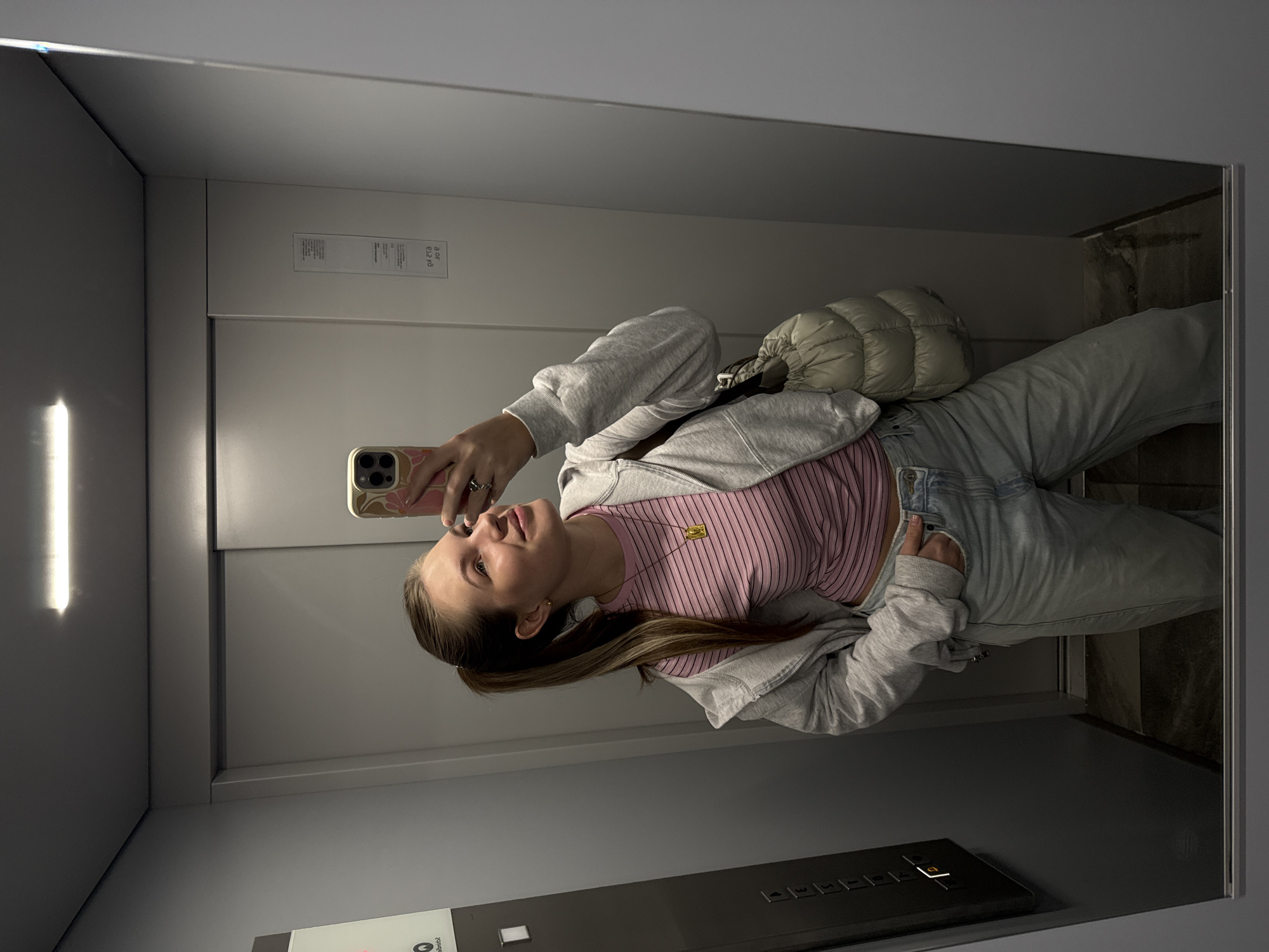 A young woman is captured in a mirror selfie inside an elevator, with the image rotated ninety degrees counter-clockwise. She smiles subtly, holding a smartphone with a patterned case up to take the picture. Her attire consists of a pink striped top, a light grey zip-up hoodie, and light blue jeans. She wears a gold necklace with a square pendant and rings on her left hand, and a light-colored, quilted shoulder bag is visible over her shoulder.
The elevator interior features reflective surfaces that act as mirrors on the walls and ceiling. A vertical fluorescent light source reflects prominently on the left side. On the right wall, a white label displays the text "B 02 / 0.12 kg". The lower part of the image shows a dark elevator control panel with floor number buttons, including '1' through '5', and a door-open icon, with one button illuminated in orange. The setting is an indoor, modern elevator, likely in Vozdovac Urban Municipality, Serbia.