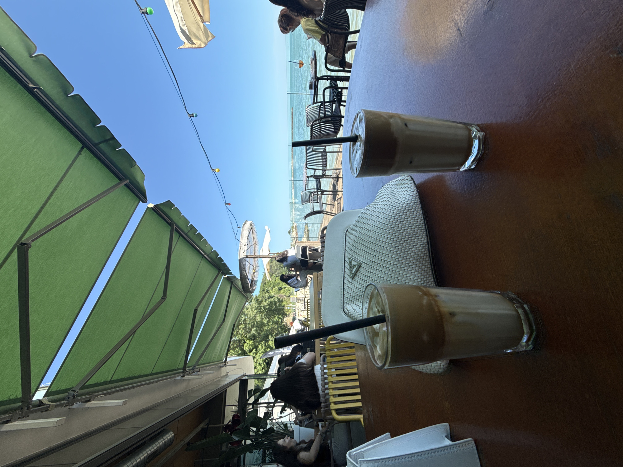 This image depicts a cafe situated on the beachfront in Varna, Bulgaria. The cafe features a green awning providing shade for outdoor seating. In the foreground, two glasses of a layered coffee drink, likely a dalgona coffee, are prominently displayed on a wooden table. A white, textured clutch bag is placed between the two glasses. In the background, several people are seen seated at tables, enjoying the sea view. The sea is calm and blue, meeting a clear sky dotted with string lights. The overall atmosphere suggests a relaxed dining experience by the coast.