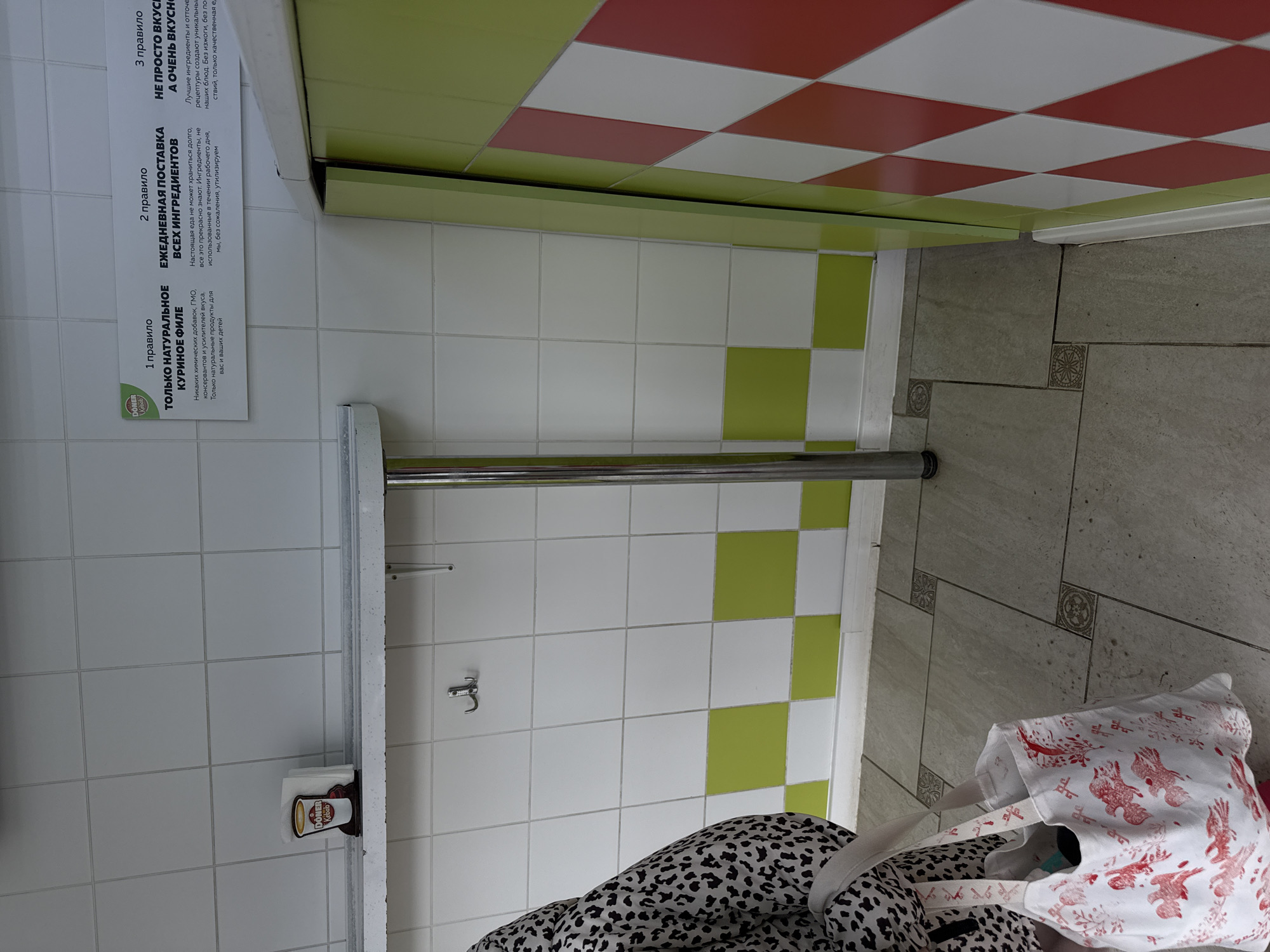 Here is a description of the image:
**Scene:** An indoor restroom, seemingly in a commercial setting.
**Location:** Likely in Perm, Russia, based on the Cyrillic script in the signage.
**Objects:**
* Tiled walls: White tiles interspersed with light green tiles.
* Tiled floor: Light gray tiles with decorative inlays.
* Ceiling: A pattern of red, white, and green tiles.
* Stainless steel grab bar.
* Toilet paper dispenser with a "Doner Kebab" branded advertisement.
* Hook.
* A sign on the wall with information in Russian, mentioning "Only Natural Chicken Fillet," daily deliveries, and taste.
* A partially visible shopping bag with a bird pattern and a black and white patterned garment.
**Subjects:** No people are visible in the image.
**Actions/Interactions:** None are directly depicted.
**Environment:** A clean, tiled restroom. The presence of the food advertisement suggests a possible cafe or restaurant.