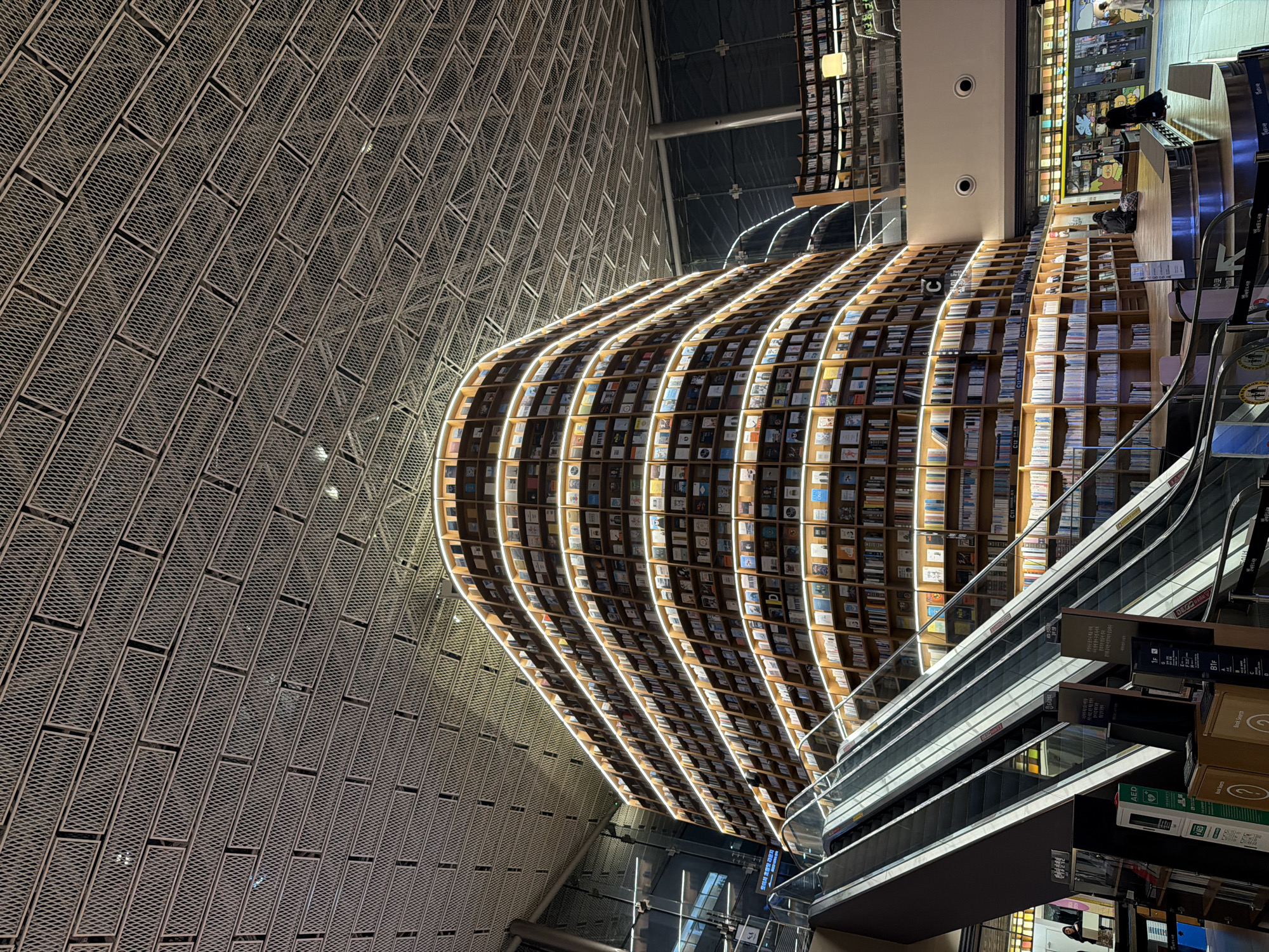The photograph depicts an interior view of the Starfield Library in the COEX Mall of Seoul, South Korea. The library is characterized by its multi-story bookshelves that curve upwards, creating a visually striking effect. The shelves are filled with books and illuminated by strip lights. An escalator is seen to the right of the bookshelves, leading to upper levels. The ceiling is high and designed with a grid-like pattern. The photograph is taken at an angle.
