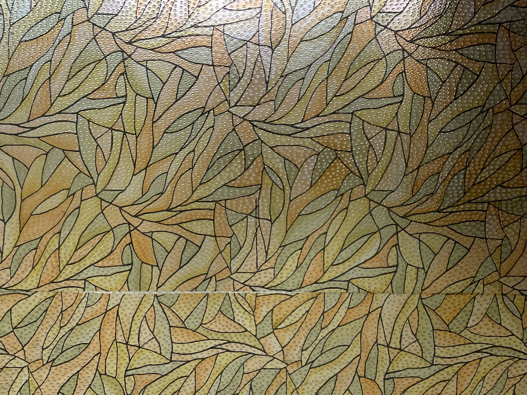 The image displays a close-up of a decorative glass panel featuring an abstract pattern resembling stylized leaves or organic shapes. The colors are a blend of warm yellows, oranges, and muted greens, with thin black lines outlining the various shapes. The glass surface has a textured, bumpy quality, as evidenced by the small raised dots visible within each colored section. There is a vertical seam running down the center of the image, suggesting the panel might be composed of multiple pieces. The lighting appears to be coming from the front and slightly to the right, creating subtle highlights and shadows that emphasize the texture of the glass. The context of the image being in Hrodna, Belarus, suggests this decorative panel could be a feature of a building's interior or exterior in that city, potentially in a historical or architecturally significant location. No people, actions, or interactions are depicted in this image.