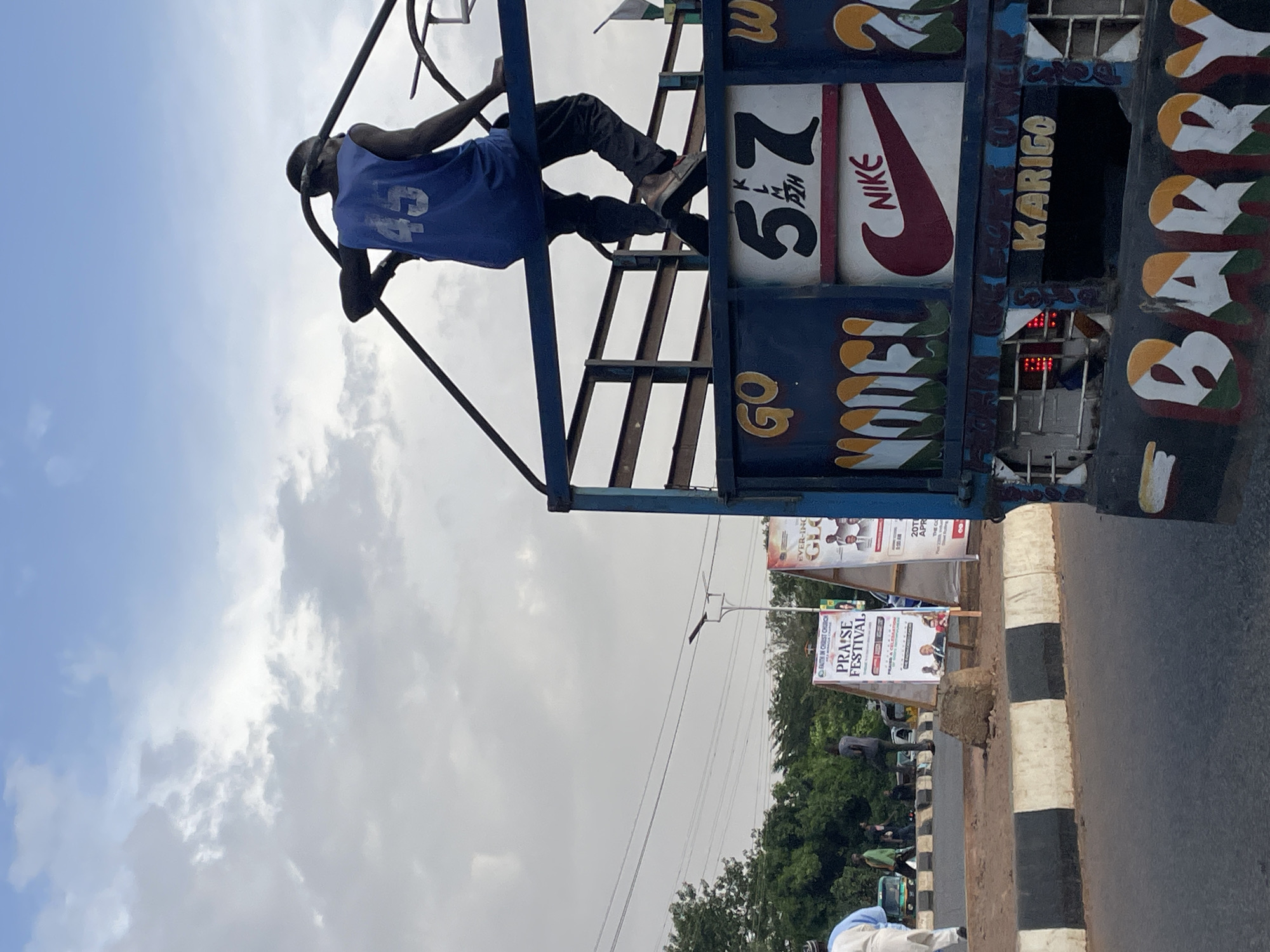 The image captures a scene in Jos, Nigeria. A man is positioned on the upper part of a vehicle adorned with vibrant, hand-painted graphics, including a depiction of the Nike logo, the word "KARIGO," and the number "57". The man is wearing a blue sleeveless jersey with the number "45" visible on the back. Below the vehicle, posters advertise a "Praise Festival" organized by the "Faith In Christ Church". The surrounding environment shows a road with a striped curb and some greenery. The sky is partly cloudy.