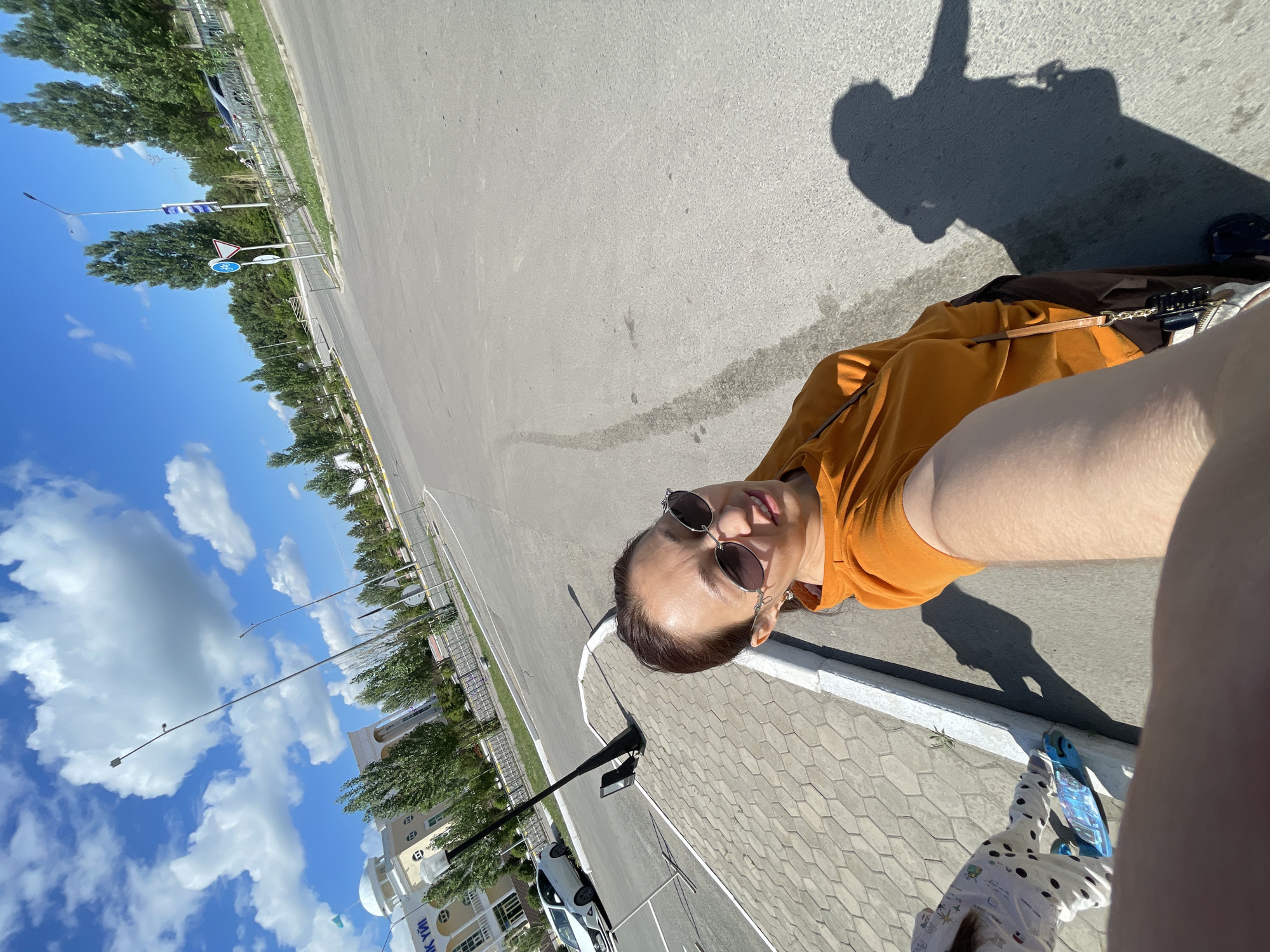 A woman wearing an orange hoodie and sunglasses is taking a selfie outdoors. She is standing on a paved sidewalk next to a road, with a building and trees in the background. A child on a blue skateboard is also visible on the sidewalk. The scene appears to be in Kokshetau, Kazakhstan, as indicated by a flag in the background and architectural style of the building. The sky is blue with scattered clouds, and there is a shadow of the woman cast on the road.