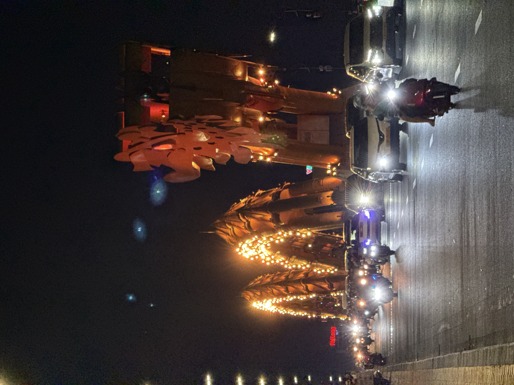 This nighttime image captures a vibrant street scene in Đà Nẵng, Vietnam. On the left side, two large, elaborately decorated structures are brightly illuminated with warm orange and yellow lights, including numerous string lights. These structures have an ornate, possibly traditional Vietnamese design, resembling either intricate buildings or large decorative floats with dragon-like or cultural motifs.
The right side of the image features a multi-lane street that appears wet and reflective, indicating recent rain. The street is populated by multiple vehicles, predominantly motorcycles and some cars, all with their headlights and taillights brightly lit, casting strong glows and reflections on the damp road surface. People are visible on or beside the motorcycles, and other individuals can be seen further back near the illuminated structures.
The time of day is clearly night, with the darkness serving as a backdrop for the various artificial light sources. The scene conveys a lively and festive atmosphere, likely during a special event, festival, or simply a bustling evening in the city. On one of the illuminated structures in the mid-ground, a red sign with white text reads "PHULONG".