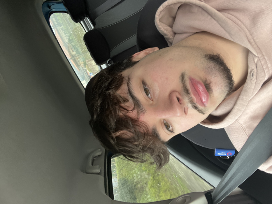 An image taken from inside a vehicle depicts a young adult male in the foreground. He has dark, wavy hair, light brown eyes, a mustache, and a goatee. He is wearing a light peach-colored hooded sweatshirt and is looking directly at the camera. A black seatbelt is visible across his chest, buckled. To the lower right of his face, a small blue cylindrical object with "SPARCO CORSE" branding and a black lanyard is visible near the seatbelt connection point. The vehicle's interior features a light grey headliner and black rear seat headrests. Through the vehicle's windows, a blurred exterior environment is visible, indicating motion. Green vegetation is prominent on the left, while the rear window shows houses with light-colored walls and tiled roofs, interspersed with more greenery and a visible utility pole or street sign. The specific location within Santa Marta de Penaguião, Portugal, is not discernible from the visible external features, but the environment suggests a residential or semi-urban road.