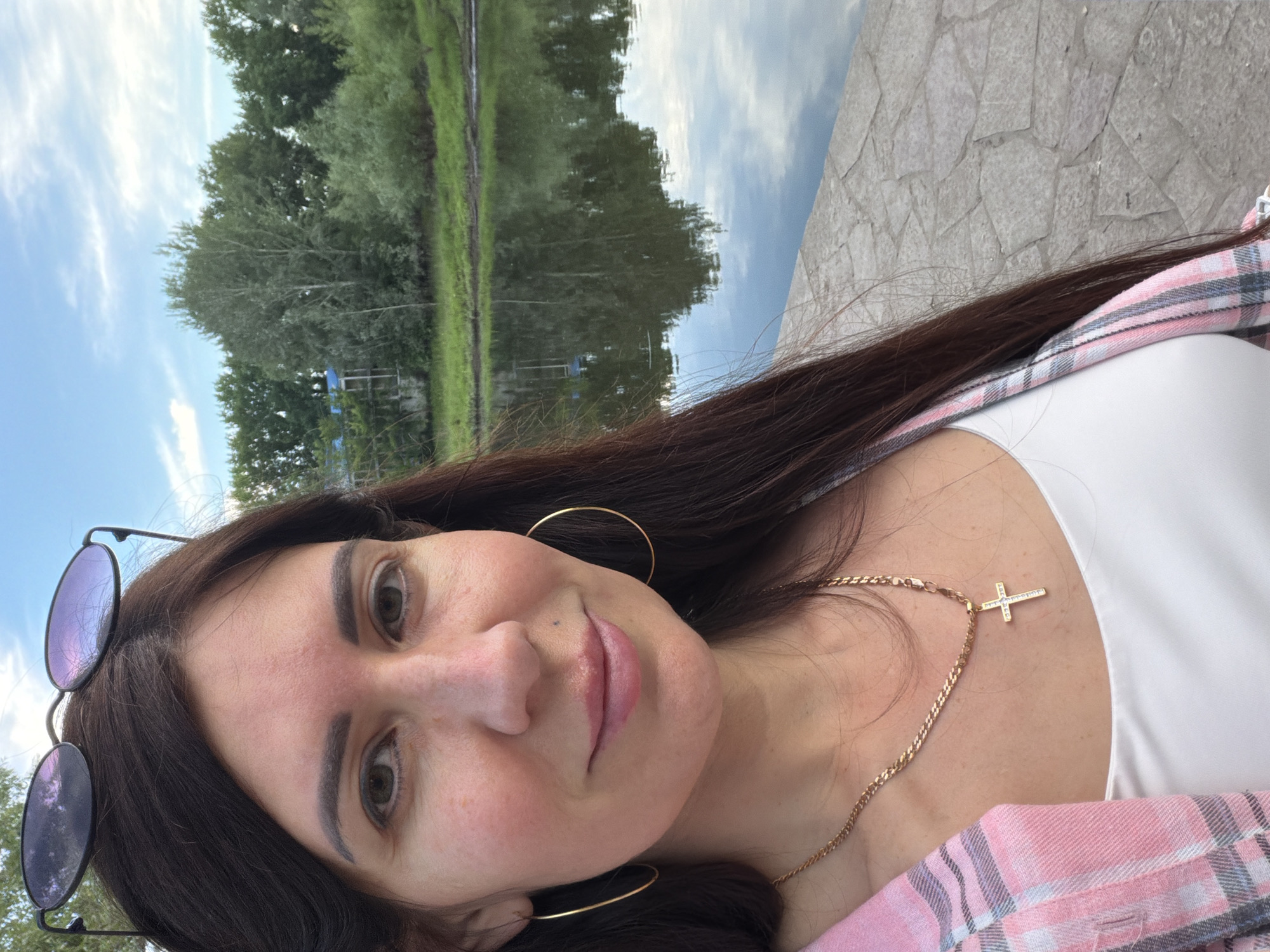 A woman is taking a selfie outdoors near a body of water. She has long, dark brown hair and is wearing sunglasses on top of her head. She is also wearing a gold chain with a cross pendant, hoop earrings, and a pink and white plaid shirt over a white top. The background shows a tranquil river or lake with trees and a cloudy blue sky reflected on the surface. The ground in the foreground is paved with cobblestones. The image was taken in Volzhskiy, Russia.
