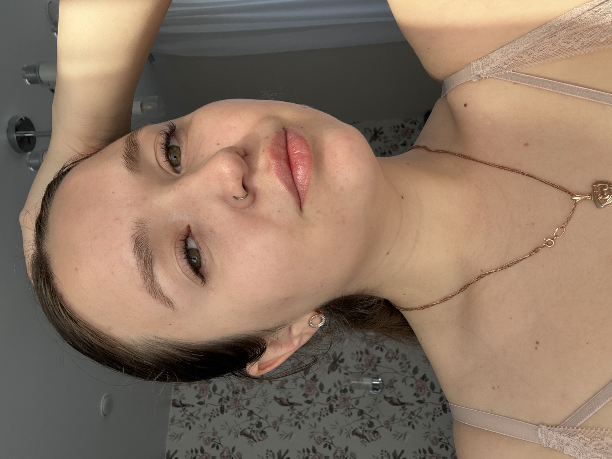 A young, fair-skinned woman is angled toward the camera. She has light brown hair, green eyes, and a nose piercing. She wears a light pink bra and a gold necklace. The background consists of a floral patterned wall and a partially visible shower head. The location cannot be determined from the image provided.