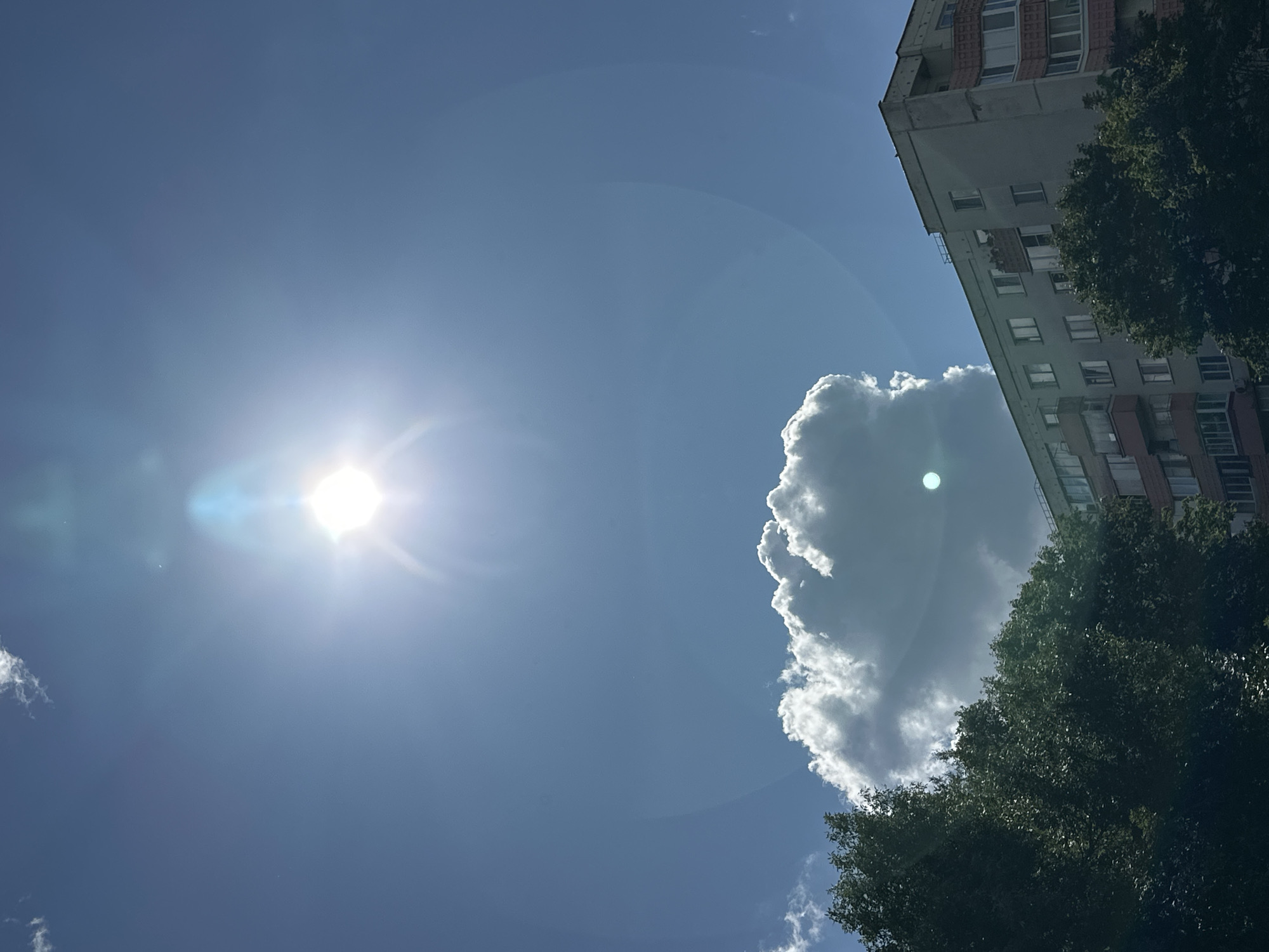 A bright sun shines in the upper center of the sky, creating a lens flare effect. Below the sun, a large, fluffy white cloud is positioned. In the lower right corner of the image is a multistory apartment building with visible windows and balconies. In the lower left and center of the image, dark green trees obscure part of the building. The sky is a clear, bright blue. This scene appears to be in an urban environment, possibly in the city of Minsk, Belarus, given the architectural style of the apartment building.