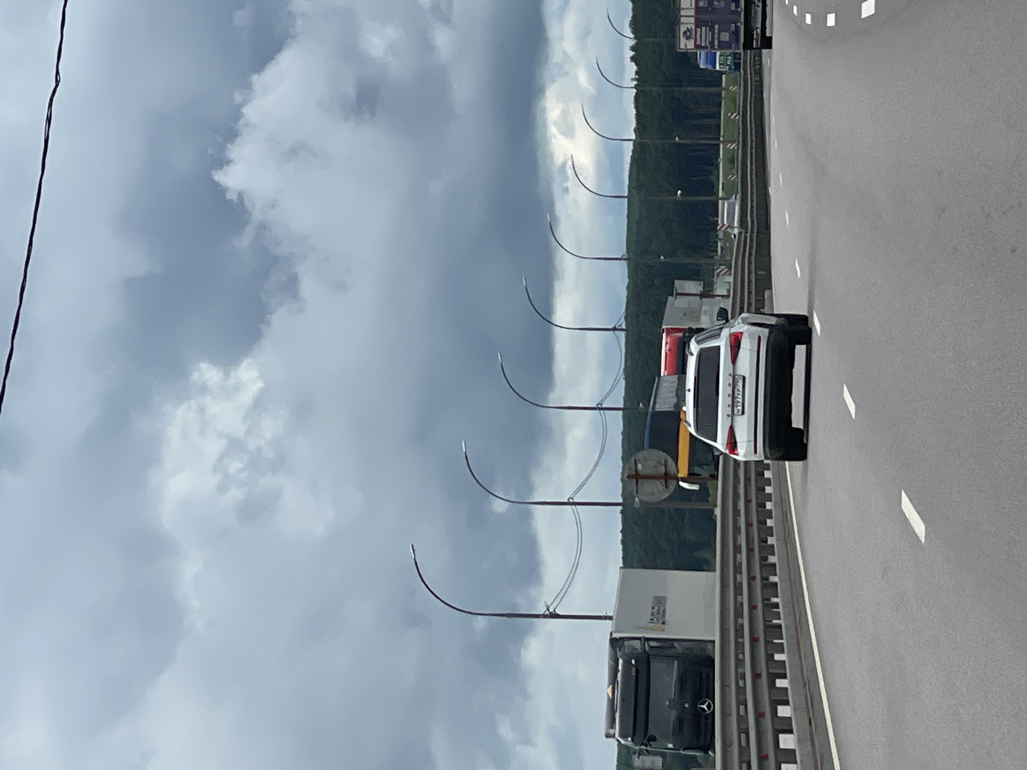 The image shows a highway in Taraskovo, Russia, under a cloudy sky. A white SUV is traveling on the road, followed by a truck. Street lights line the highway. Trees are visible in the background.