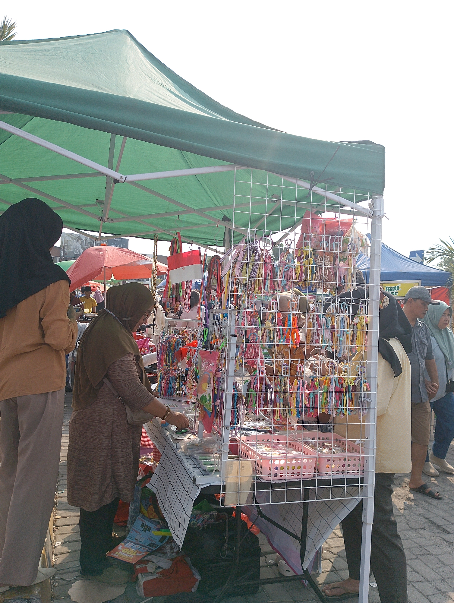 A street market is depicted in Karawang, Indonesia, with a focus on a stall selling various colorful trinkets. The stall is covered by a green awning, and displays a large assortment of small, brightly colored items hanging on a white wire grid. Two women are actively engaged at the stall. One woman, wearing a brown hijab and a long brown patterned tunic, is examining the items. Another woman, wearing a black hijab and a tan long-sleeved shirt, stands to her left, facing away from the camera. Behind them, other market stalls and people are visible. One stall has a red and white flag, possibly the Indonesian flag. The ground is paved with stones, and the scene appears to be outdoors under daylight. Several pink plastic baskets filled with more trinkets are also visible on the stall.