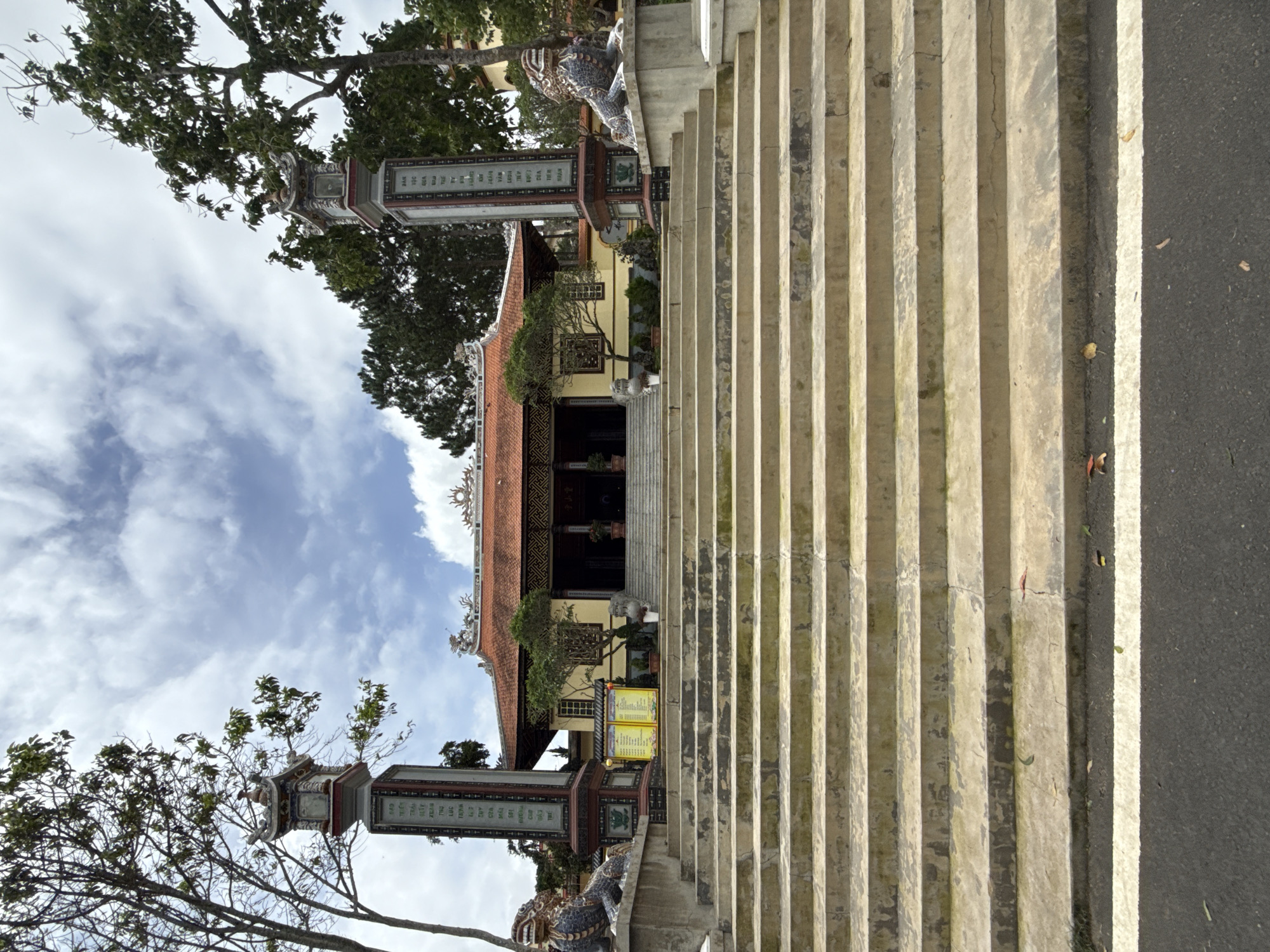 The image shows the wide, worn stone steps leading up to a Buddhist temple in Da Lat, Vietnam. The temple itself has a traditional Vietnamese architectural style with a red tiled roof. Flanking the stairway on either side are large, ornate pillars topped with decorative elements and statues. To the left and right of the base of the pillars, there are stone guardian lions. The steps are in the foreground, with the temple and its surroundings in the background, under a cloudy sky. The overall scene conveys a sense of age and spiritual significance.