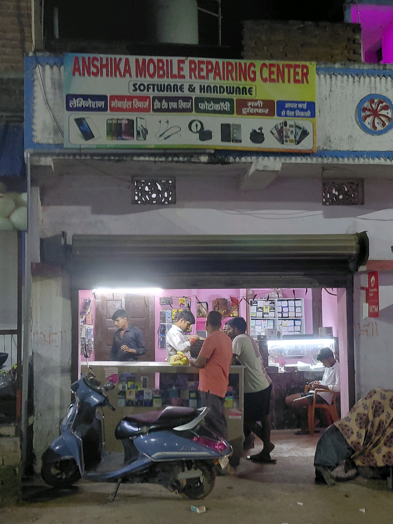 The image depicts the exterior of "ANSHIKA MOBILE REPAIRING CENTER" in Patna, India. The shop's facade features a prominent sign with the business name and services like "SOFTWARE & HARDWARE," "LAMINATION," "MOBILE RECHARGE," "PHOTOCOPY," "MONEY TRANSFER," and "AADHAAR CARD SE PANIKALRI." Below the main sign, smaller icons represent various services and products, including mobile phones, earphones, and computer peripherals.
The shop's interior is illuminated, revealing shelves stocked with mobile accessories and phones. Three men are visible inside the shop: one in a blue shirt standing behind a counter, another in a white shirt leaning forward, and a third in a white shirt seated on a chair, looking at his mobile phone. A blue scooter is parked outside the shop. The scene appears to be in a commercial area, with the shop situated on a street. The overall ambiance suggests an active business environment at night.