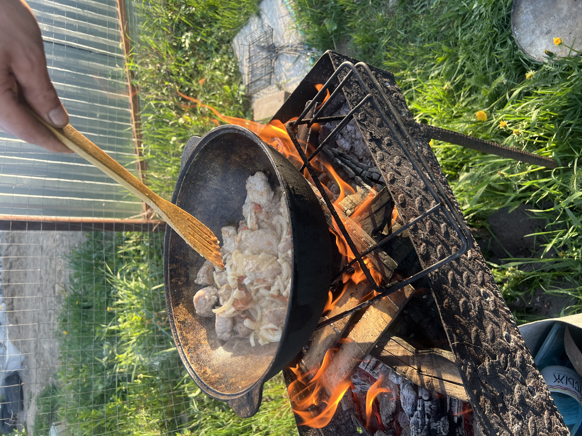 A cast-iron pot is situated atop a grill with burning wood. The pot contains meat and onions being stirred by a person's hand holding a wooden spatula. The scene is set outdoors on a grassy area in Chaikovsky, Russia. A greenhouse structure is partially visible in the background, alongside assorted tools and items scattered on the grass.
