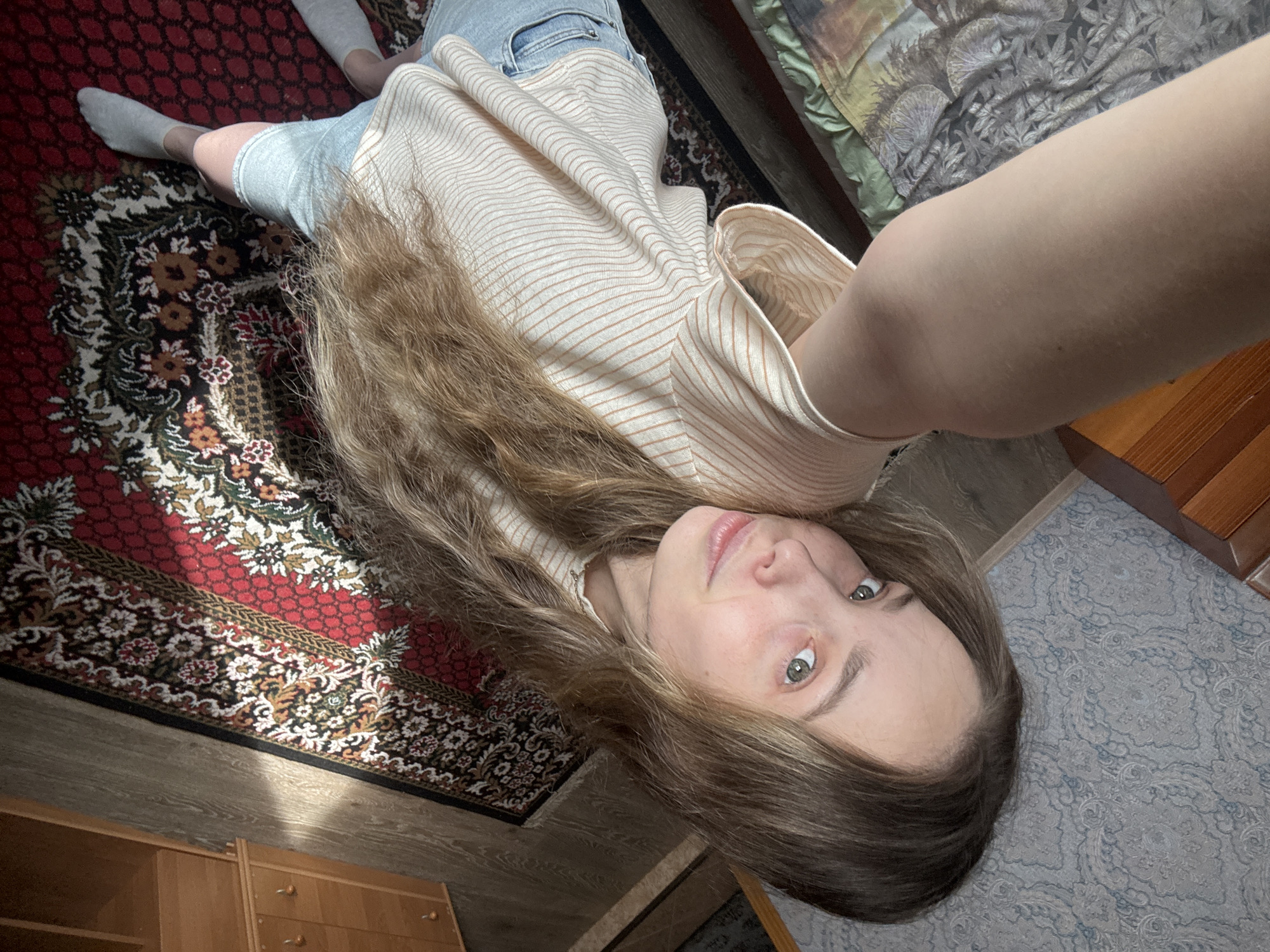 A young woman with long, light brown hair and light green eyes is captured in a high-angle, slightly blurred selfie. She is wearing a striped shirt and denim shorts. Her left arm is extended upwards, holding the camera. She is indoors, standing on a patterned rug in what appears to be a bedroom. Sunlight streams in from the right, casting shadows. To the left of the woman, a bed with a patterned comforter is partially visible. To the right, a wooden dresser with drawers and knobs is present. The floor is wooden. The specific location within Prikomsky, Russia, cannot be determined from the visual information provided.