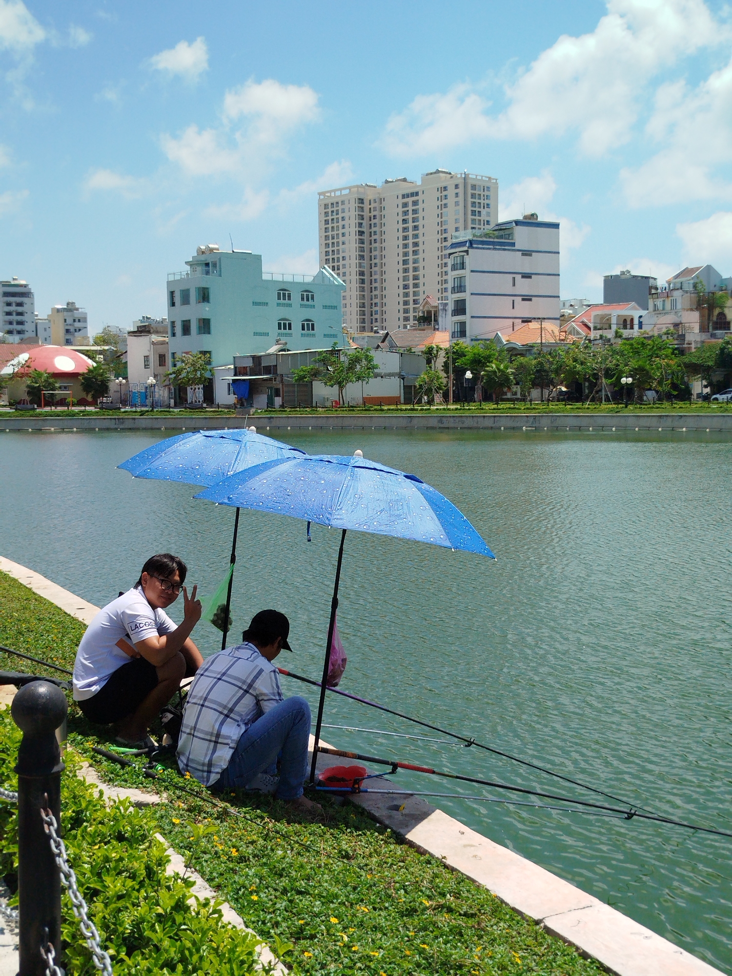 Two men are fishing under a blue umbrella at the edge of a lake in Ho Chi Minh City, Vietnam. One man is facing the camera, wearing a white Lacoste t-shirt and giving a peace sign. The other man is wearing a blue and white plaid shirt and blue jeans, with his back to the camera. Fishing rods are lined up along the edge of the lake. In the background, there are several buildings of varying heights, and some trees. The sky is blue with scattered clouds. A black metal fence and green bushes are in the foreground.