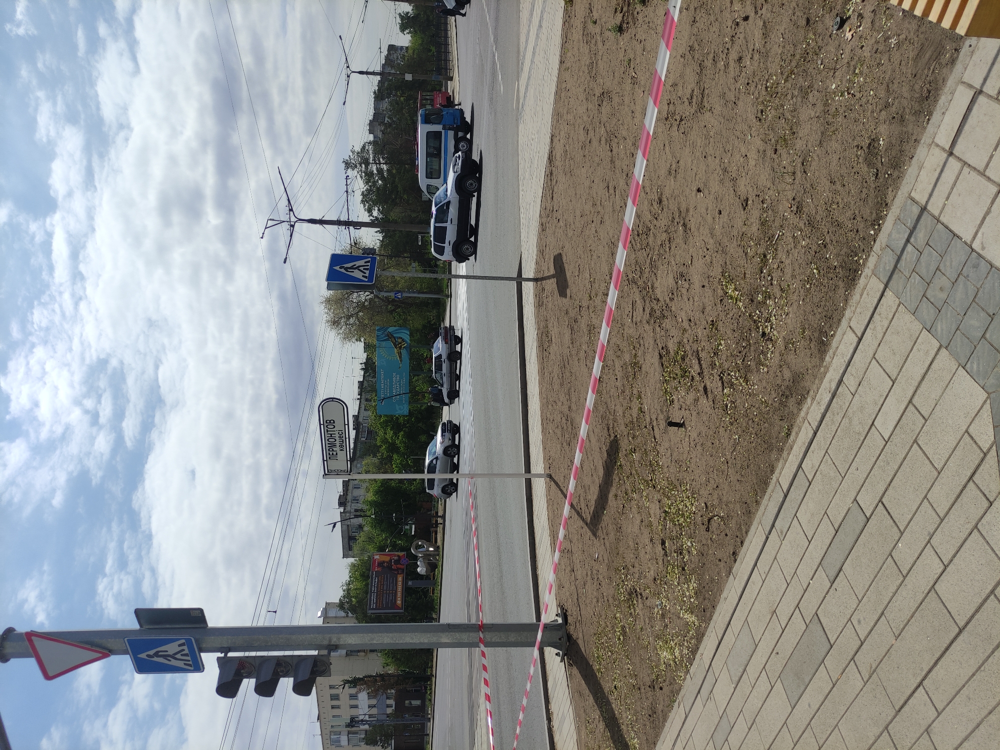 The image shows a street scene in Pavlodar, Kazakhstan. A street sign reads "Lermontov Street" in both Russian and Kazakh. There are several vehicles on the road including cars and a white van. Pedestrian crossing signs are visible. A construction zone is indicated by a red and white striped barrier tape. The weather is partly cloudy. The ground surface next to the sidewalk is dirt with some vegetation. The sidewalk is made of gray and white paving stones. Overhead are power lines and traffic signals.