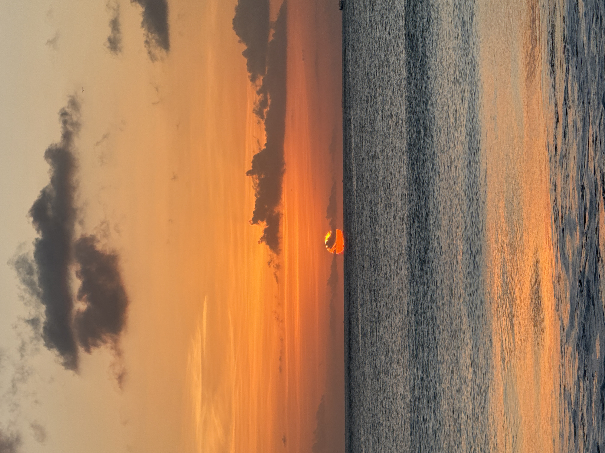 A serene ocean sunset is captured in a vertical photograph from the coastline of Kuta Selatan, Indonesia. The sky is awash in vibrant hues of orange and yellow, with streaks of lighter orange painting the horizon. Dark, amorphous clouds are scattered across the upper portion of the sky, some appearing heavy and deep gray, while others are lighter and more diffused. A sliver of the sun, a brilliant orange orb, is partially obscured by clouds near the horizon, casting a warm glow.
The ocean occupies the lower two-thirds of the frame. The water is a deep, dark blue, reflecting the warm tones of the sky in a band of shimmering orange and yellow light across its surface. Gentle waves are visible in the foreground, with white foam cresting on their edges. In the distance, a dark silhouette of what appears to be a boat is visible on the right side of the horizon. The overall atmosphere is one of tranquility and natural beauty.