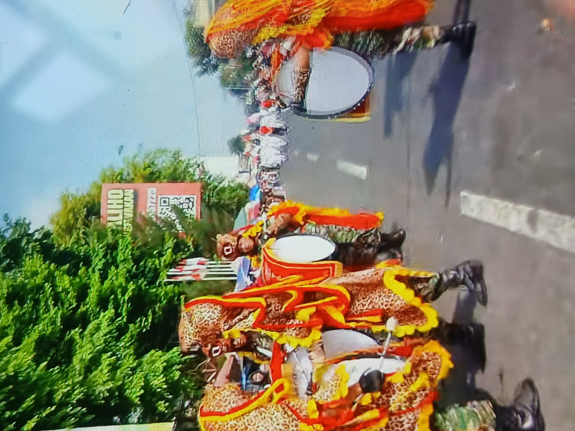 A parade is taking place on a paved street in what appears to be Setia Bakti, Indonesia. In the foreground, three individuals are dressed in elaborate costumes that resemble leopards, with red and yellow trim. They are wearing camouflage pants and black boots. Two of them are carrying large bass drums. To the left, a banner with Indonesian text and a QR code is visible. In the background, a crowd of people, some in white outfits, are participating in the parade. Trees line the side of the street, and the sky is clear and blue. The overall atmosphere suggests a festive cultural event.