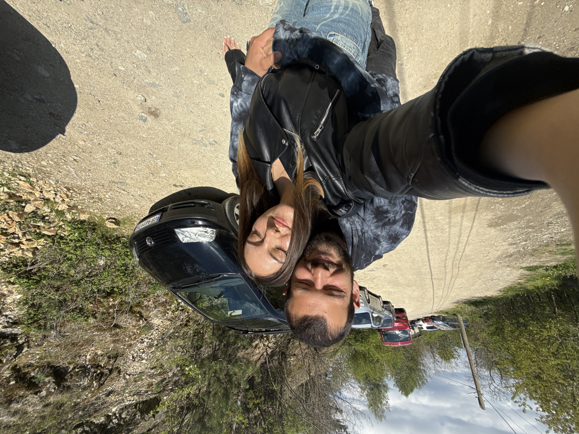A man and a woman are posing for a selfie on a dirt road. They are standing in front of a black car, partially visible. In the background, there are several other cars parked along the road. The scene appears to be in a mountainous or wooded area. The setting is near Ovchartsi, Bulgaria, as the car's license plate (EB 9496 BS) is a Bulgarian one. The woman has brown hair and is wearing a black leather jacket over a tie-dye shirt. The man is wearing a black leather jacket. They both have their eyes closed. The photo is taken from an overhead perspective.