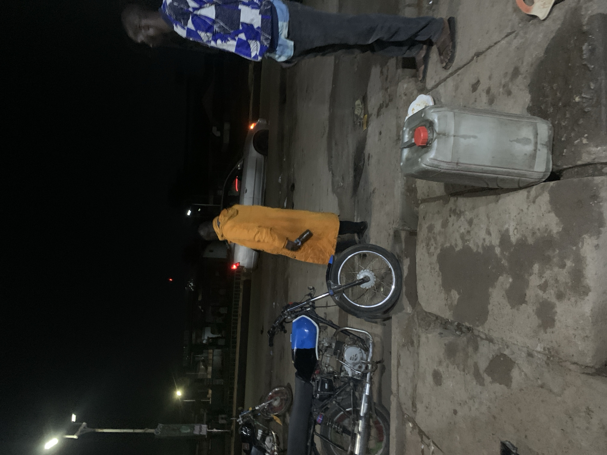 This nighttime image depicts an outdoor scene, likely along a street or roadside in Gura Top, Nigeria. The environment is dim, relying on artificial light sources such as distant vehicle lights and streetlights.
Two individuals are visible. In the upper part of the frame, a man with dark skin wears a blue and white patterned shirt, dark trousers, and sandals. He is standing on a concrete surface and appears to be looking down. Below him, another person is dressed in a distinctive bright orange or yellow full-body garment and dark attire, positioned next to a motorcycle. Their face is not clearly visible.
The scene features at least two motorcycles. One prominently displayed in the midground has a blue fuel tank, a black seat, and visible engine components and a spoked front wheel. A second, darker motorcycle is partially visible behind it. A large, grey plastic jerrycan with a red cap rests on the concrete ground near the first man's feet, suggesting it holds fuel or water. The ground itself is a stained and worn concrete or paved surface.
The arrangement of the jerrycan, motorcycles, and individuals implies an activity related to fuel, possibly a roadside refueling stop or an informal fuel sale. In the blurred background, the presence of vehicle lights suggests a functioning road or parking area. No visible text is present in the image. The overall impression is one of a routine, functional interaction occurring under low-light conditions.