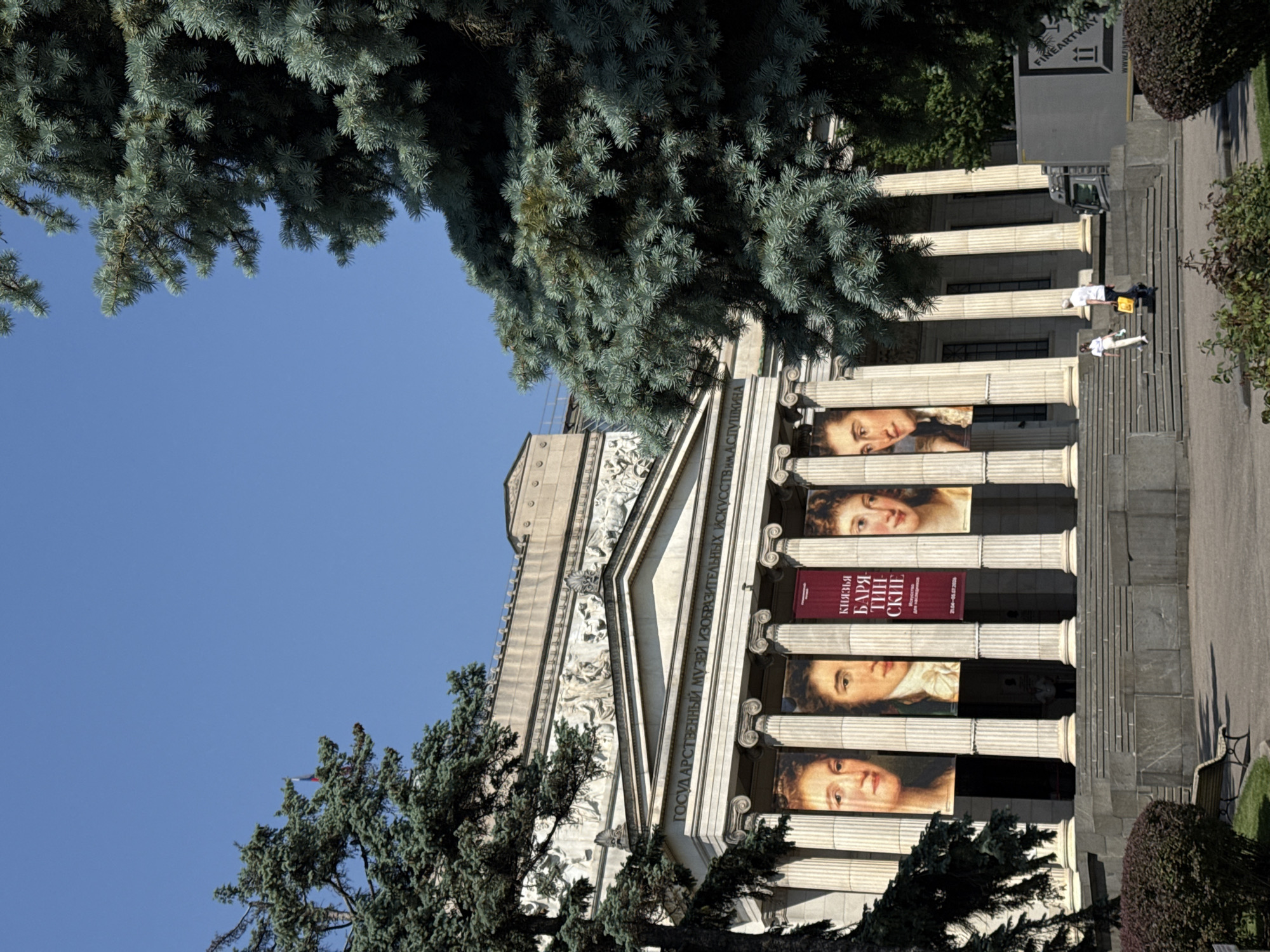 The image captures the facade of the Pushkin State Museum of Fine Arts in Moscow, Russia. The classical architecture is marked by rows of Doric columns, adorned with banners showcasing portraits. In the foreground, trees frame the scene, contrasting with the building's stone structure. Two individuals are visible on the museum's steps, lying prone.
