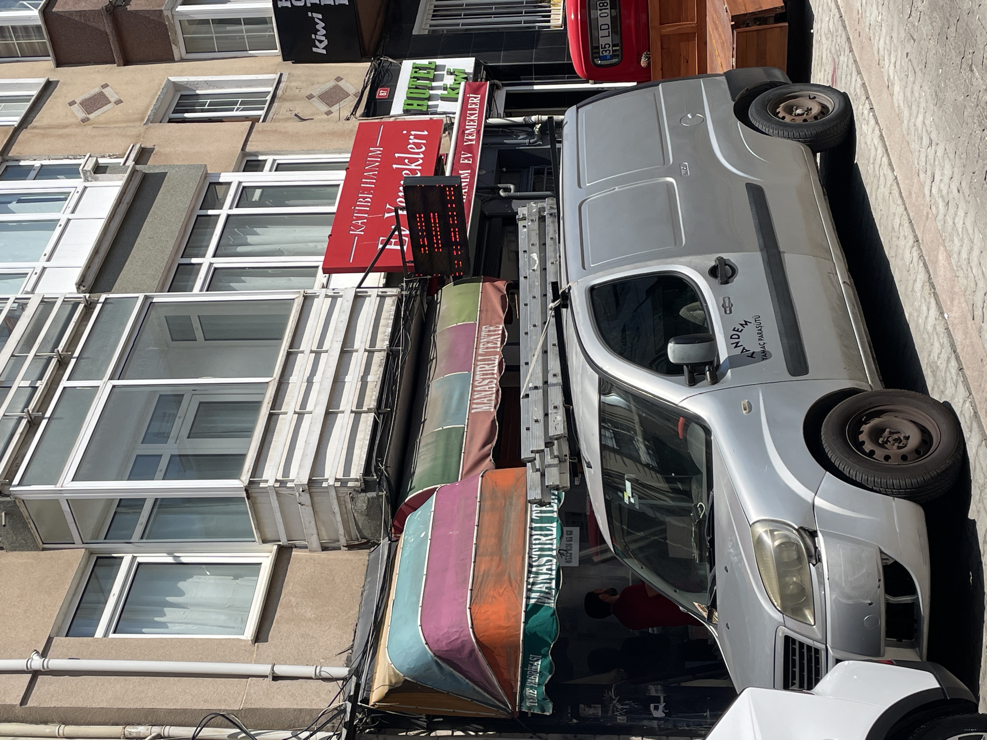 The image depicts a street scene in Kadıköy, Turkey. A silver panel van with ladders on its roof is parked in the foreground, partially obscuring a white vehicle to its left and a red truck to its right. The van has text on its side reading "TANDEM YAMAÇ PARAŞÜTÜ".
Behind the van, a building displays several awnings. The leftmost awning is multi-colored, featuring bands of teal, purple, and orange. To its right, another awning is green and brown, and a sign below it reads "MANASTIRLI TE..." with a phone number "0543 336 65 68". Further to the right, a red sign advertises "KATİBE HANIM EV YEMEKLERİ" (Katibe Lady Home Cooking). Above this sign, a smaller green sign reads "HOTEL Kiwi".
The building has multiple floors with windows, some with white frames and others with darker frames. Balconies are visible on some of the upper floors. The street is paved with cobblestones. The overall impression is of a commercial street with shops and services.