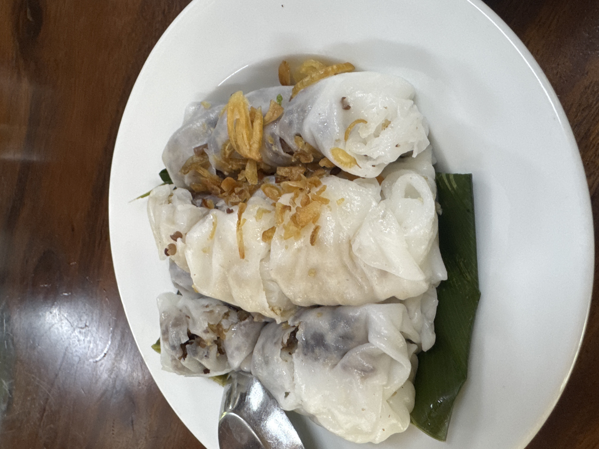 The image depicts a plate of Vietnamese steamed rice rolls, known as Banh Cuon, placed on a dark wooden table. The location is consistent with a restaurant or eatery in the city of Hà Giang 1 Ward, Vietnam, as suggested by the dish's regional origin.
The rolls appear translucent and are filled with a mixture that might contain ground pork and wood ear mushrooms. They are topped with fried shallots or garlic for added flavor and texture. A piece of green banana leaf is visible beneath the rolls, possibly used as a serving base. A metallic spoon is also visible next to the food.