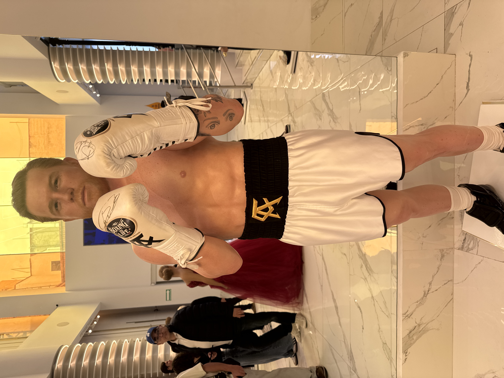 The image shows a wax figure of boxer Canelo Alvarez in a boxing pose, wearing white boxing gloves, white shorts with a black waistband bearing a gold design, and white socks with black shoes. The figure stands in front of a large mirror, which reflects lighting fixtures, a wall panel, and lettering on the mirror's surface. Reflected in the mirror are other people, including one in a red dress. Additional people are visible in the background, one wearing a blue baseball cap. The floor has a marble pattern. Based on the person in the red dress being at an unveiling event for the boxer's wax figure, the location may be a museum or public space in Mexico City, Mexico.