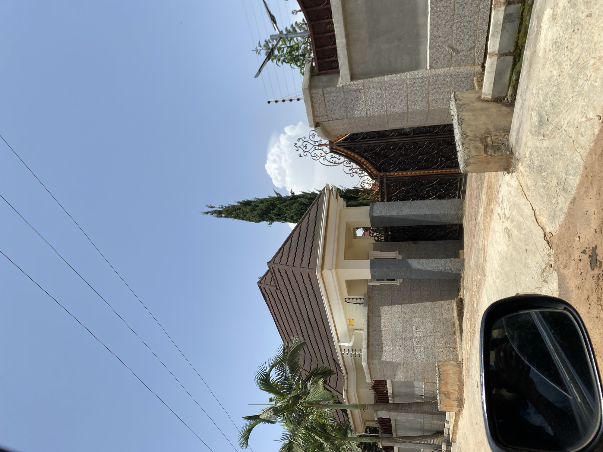 The image shows a street view of a modern house in Gura Top, Nigeria. The house features a brown tiled roof, light-colored walls with stone accents, and tall, slender pillars at the entrance. A large, ornate black metal gate is visible, leading into the property. In the foreground, a side-view mirror of a vehicle is reflected, suggesting the photo was taken from a car. A palm tree with characteristic fronds is planted near the house. The sky is clear blue with a few fluffy white clouds, indicating a sunny day. Electrical wires stretch across the sky. The scene appears to be a residential area in the daytime.