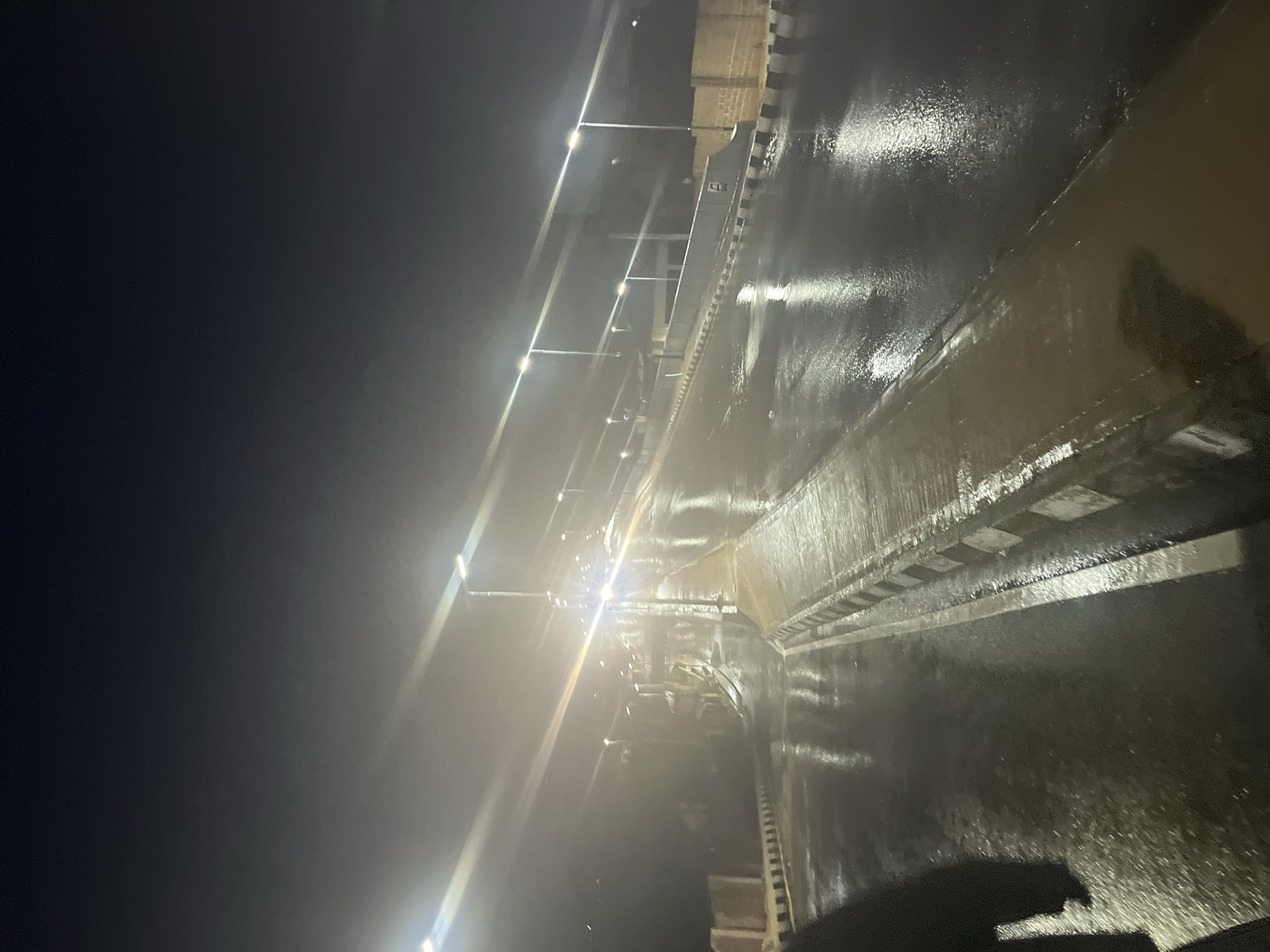The image depicts a wet road at night, likely in Jos, Nigeria, illuminated by streetlights. The road surface is reflective due to recent rainfall, showing streaks of light from the lamps. The area appears to be an elevated roadway or bridge, with a concrete barrier on the right side marked with black and white stripes. Beyond the barrier, a wall constructed of light-colored bricks is visible. On the left side of the road, a similar striped barrier is present, and further in the background, there are indications of structures or terrain. The sky is dark, characteristic of nighttime. A portion of a person's dark clothing is visible in the lower left corner of the frame, suggesting the perspective of someone standing on the side of the road or walking. The overall atmosphere is somber and damp.