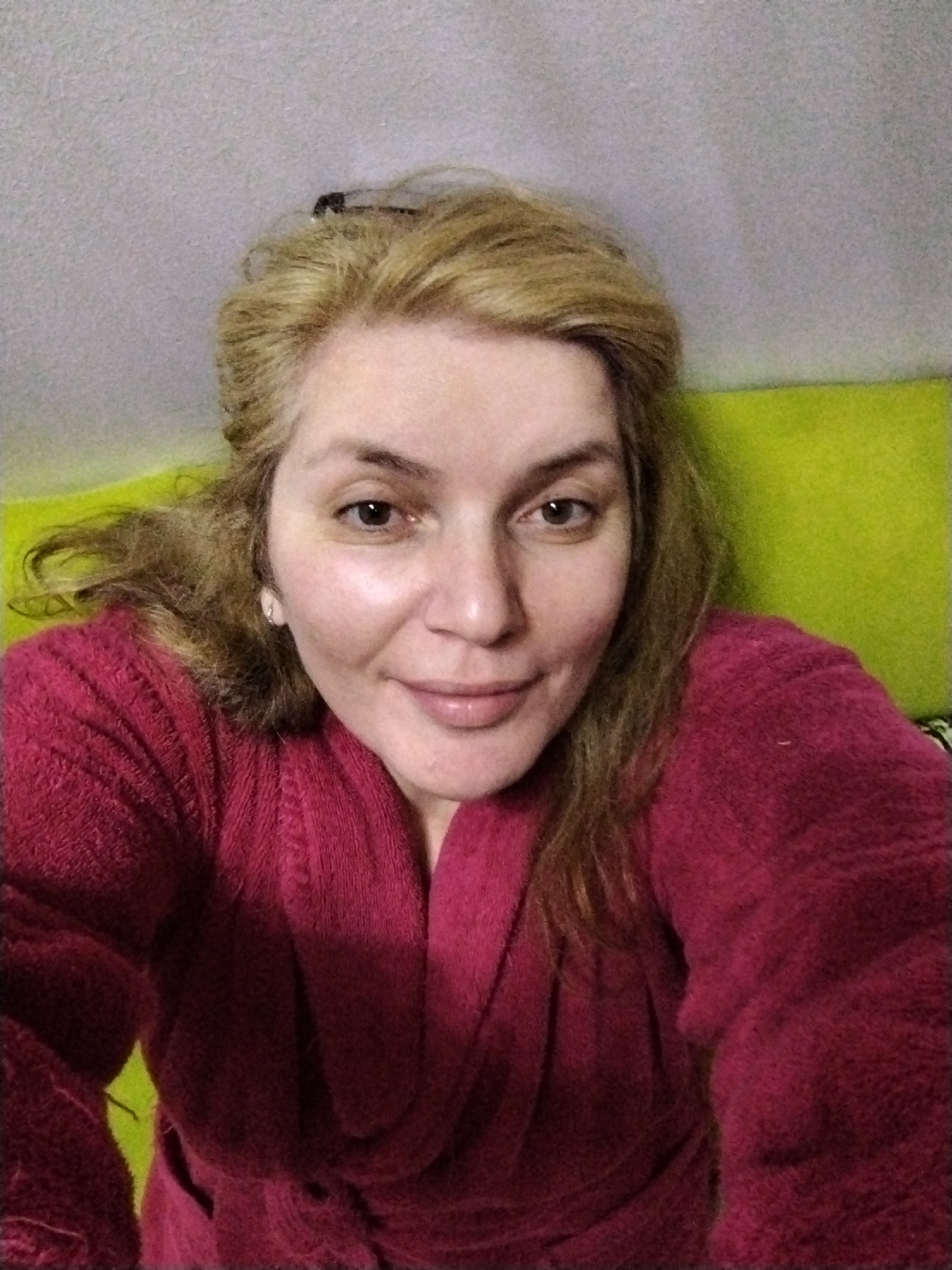 A woman is centered in the frame wearing a red, possibly terry cloth, robe. Her hair is blonde and appears to be partially pulled back. She is smiling slightly and looking directly at the camera. The background consists of a gray wall and a green surface, possibly a piece of furniture, behind her. The lighting appears to be indoor and relatively soft. There is no evidence to suggest the location is in Al Mas'oudi, United Arab Emirates, or any urban setting.