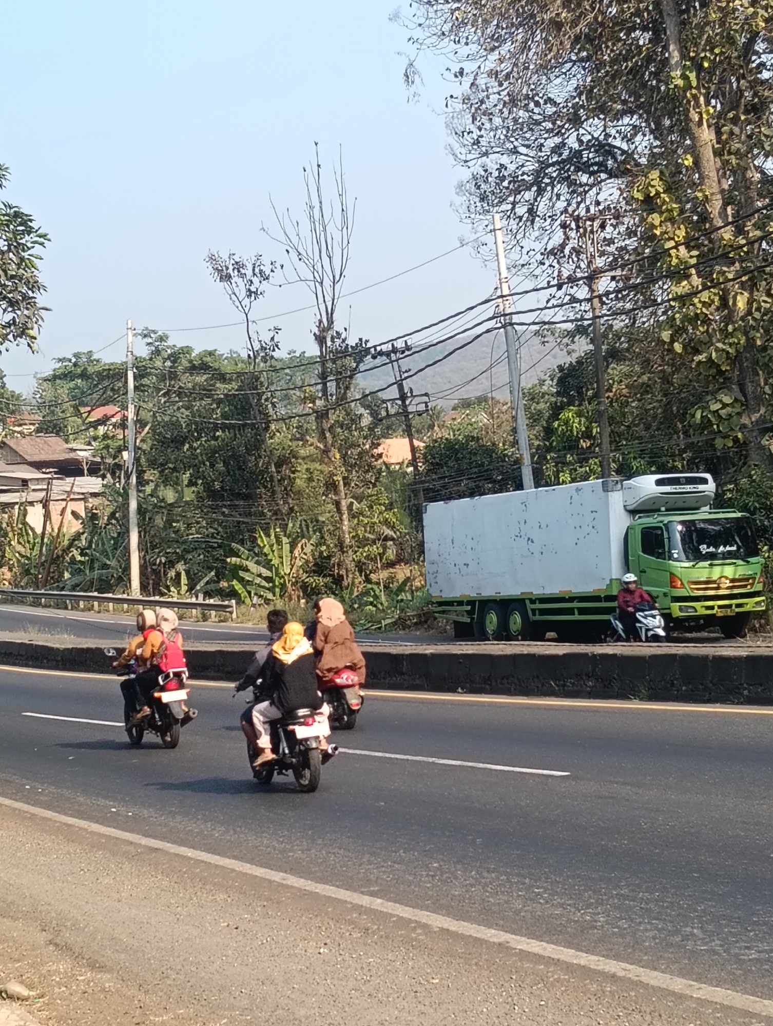 On a sunny day, a green refrigerated truck is parked on the side of a road. A person wearing a red jacket and white helmet is sitting on a motorcycle in front of the truck. In the background, there are trees, power lines, and houses. Three motorcycles with riders are traveling on the road in the foreground. The first motorcycle has two people on it, both wearing headscarves. The second motorcycle has one person wearing a yellow headscarf. The third motorcycle has one person wearing a brown headscarf. The road appears to be in Adinuso, Indonesia, given the vegetation and architectural style of the houses.
