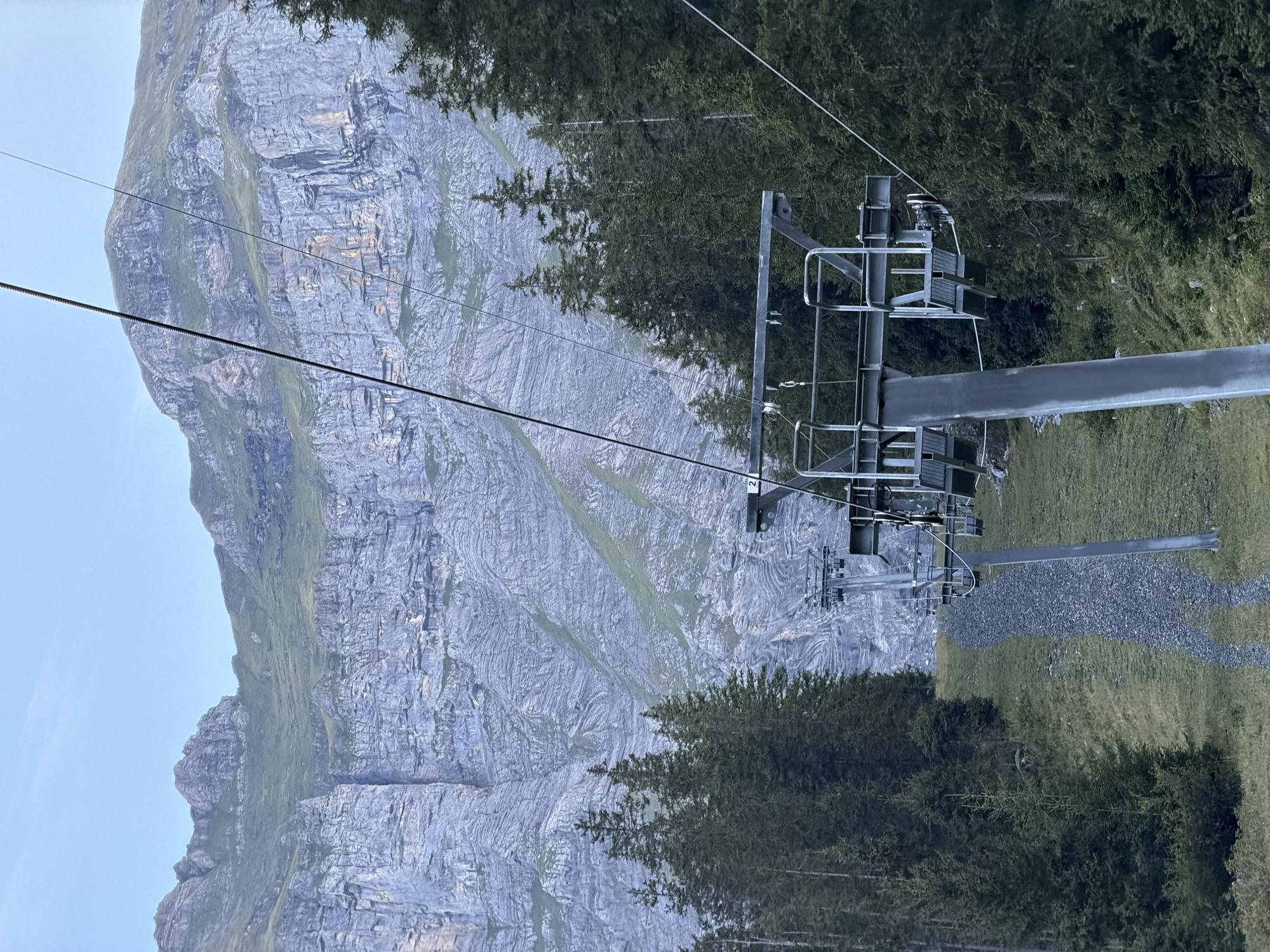 The image depicts a ski lift on a steep, grassy slope in Kandersteg, Switzerland. The ski lift consists of metal poles and chairs suspended from a cable that stretches upwards towards a rugged, rocky mountain range. Dense evergreen trees flank the slope on both sides, with a narrow, gravel path running alongside the ski lift. The sky is clear and pale blue, suggesting it is either early morning or late evening. The mountain in the background is imposing, with sheer rock faces and patches of green vegetation. No people are visible in the frame, and the ski lift appears to be stationary.