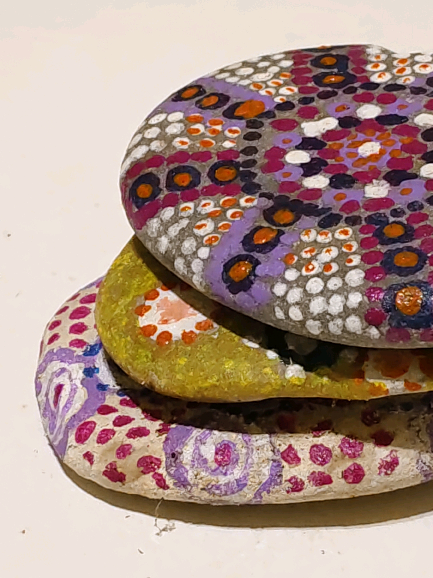 The image shows a stack of three painted stones. The top stone is decorated with a pattern of purple, pink, white, and orange dots, some of which are arranged in flower-like shapes with dark centers. The middle stone is a solid yellow with a few scattered orange and white dots, as well as a pinkish area. The bottom stone has a white base decorated with purple swirling patterns and scattered pink dots. The stones are resting on a light-colored surface, likely a table. The lighting is soft, coming from the top left. The image does not provide any information about the location in Rostov-on-Don, Russia, or any people, actions, or interactions.